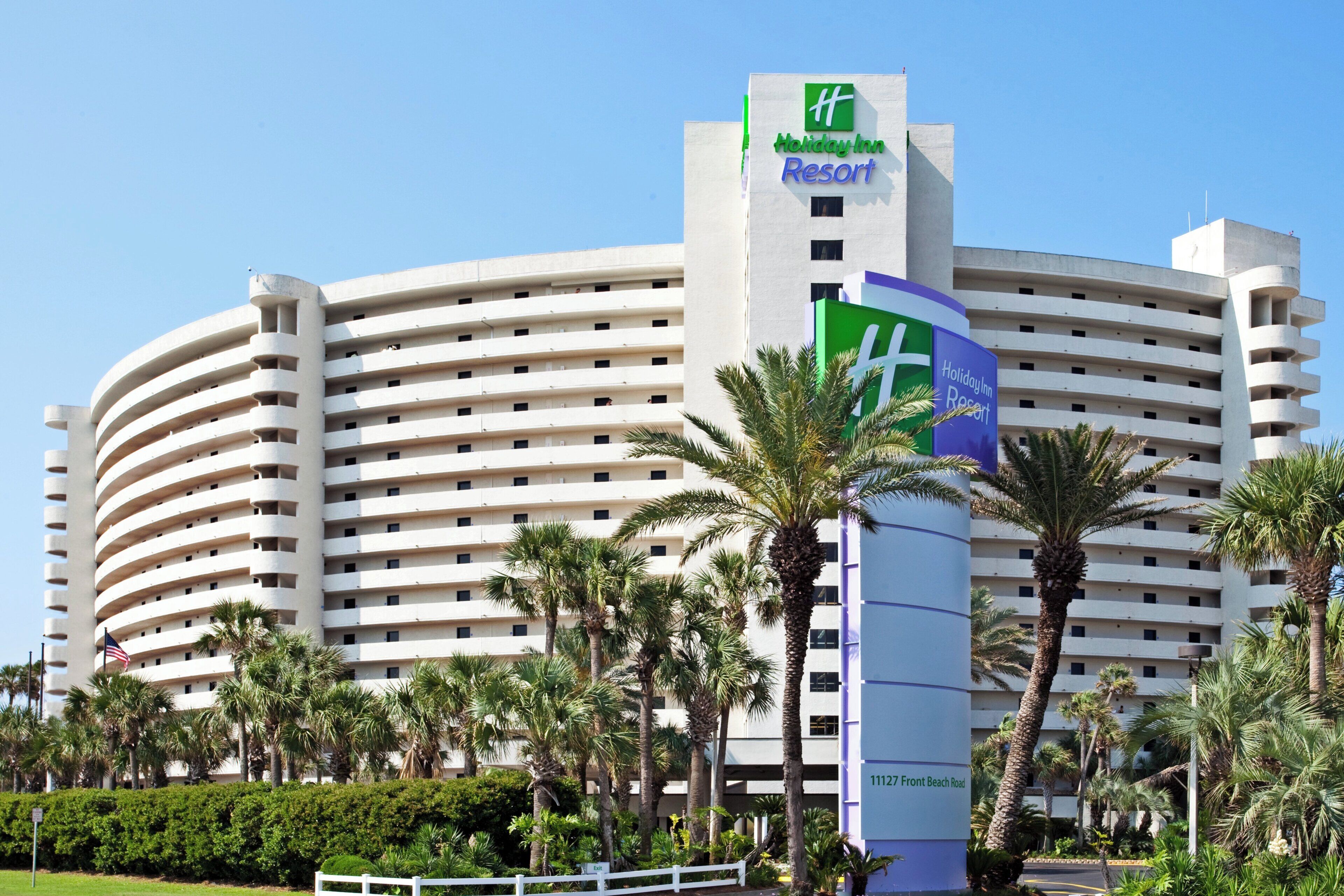 A large, multi-story Holiday Inn Resort building with a curved facade, surrounded by palm trees and a well-maintained lawn.