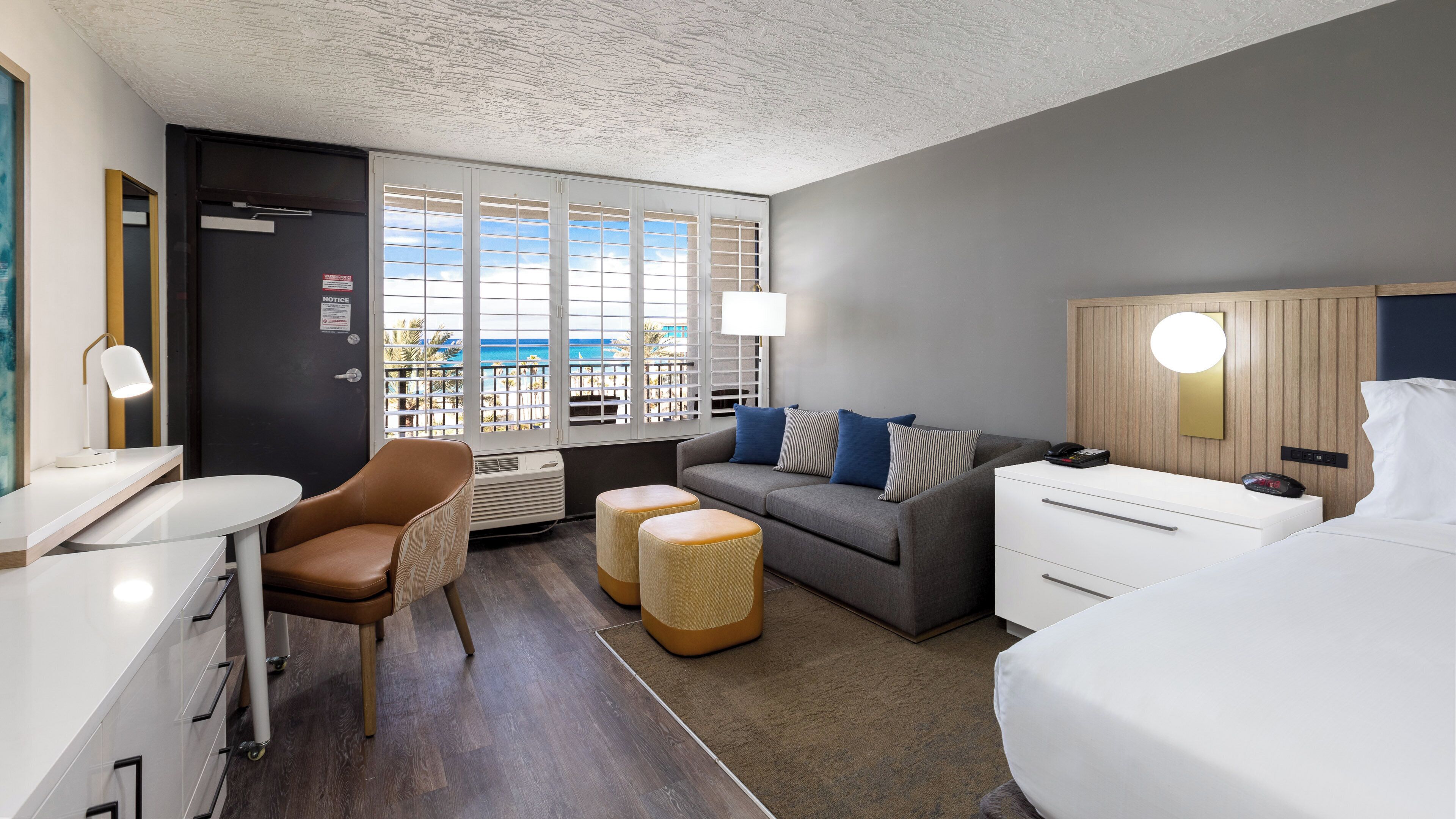 Una habitación de hotel moderna con un sofá gris, una cama, un escritorio y vista al océano a través de amplios ventanales.