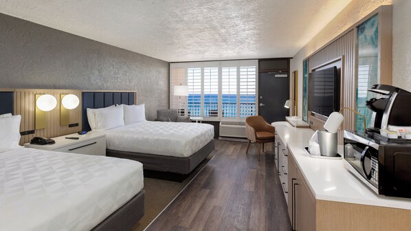Habitación de hotel con dos camas, un escritorio con cafetera, una silla y un ventanal con vista al océano.