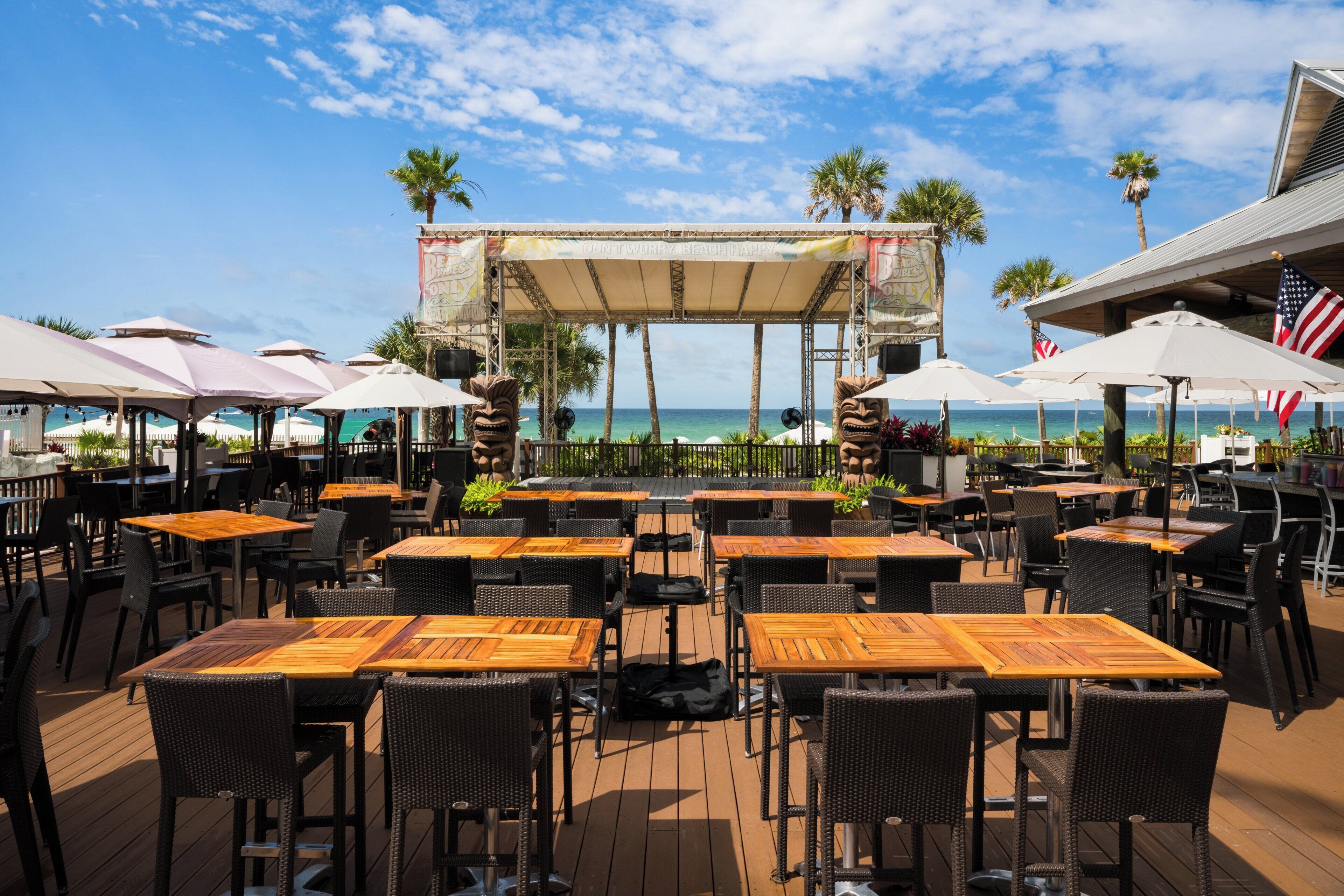 An outdoor dining area with tables and chairs, a stage with a backdrop, and a view of the ocean.