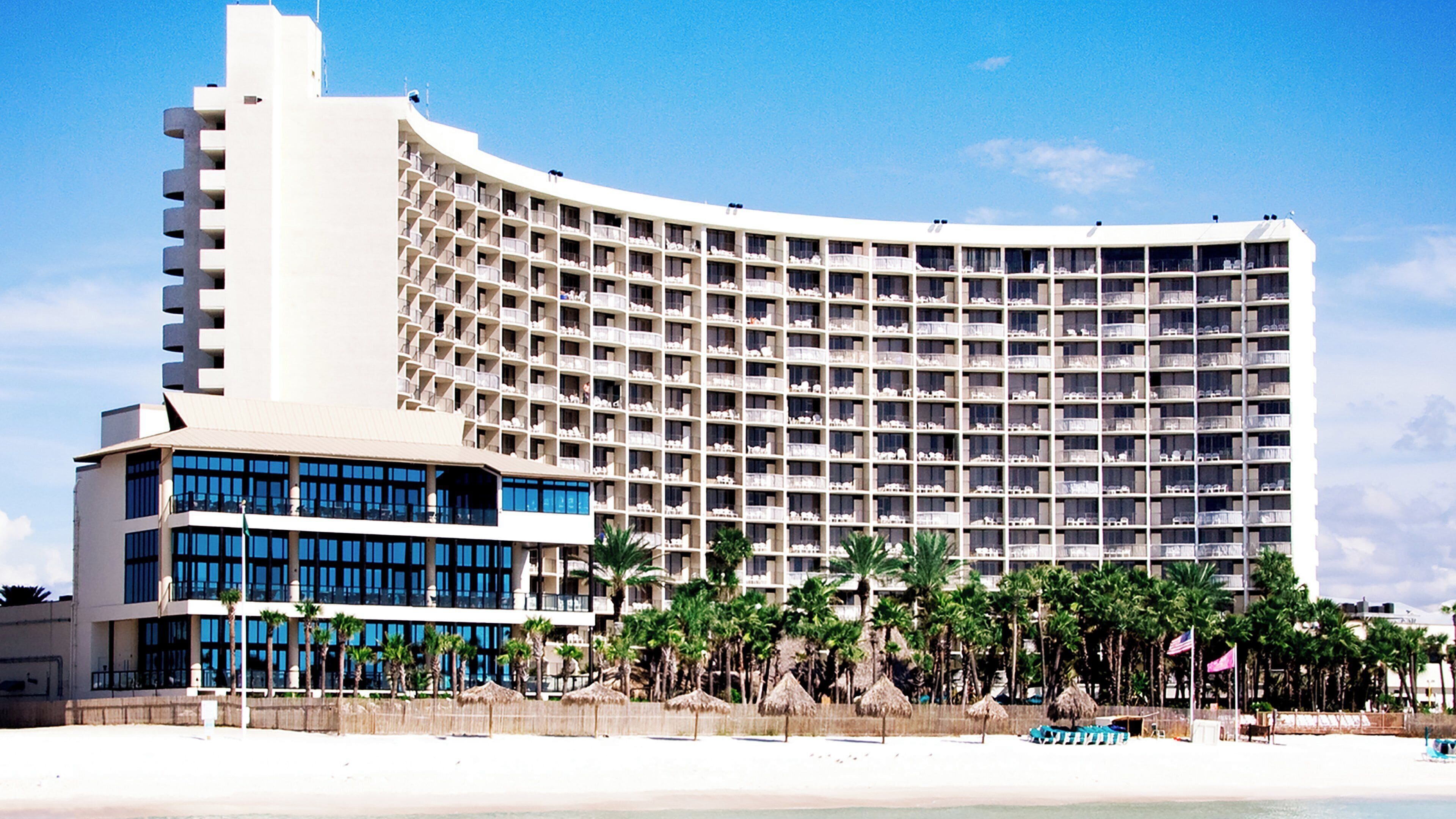 A large hotel with multiple floors and balconies, located near a beach with palm trees.