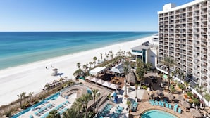 Un resort con piscina frente a la playa, palmeras y un edificio blanco con balcones.
