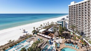 Un resort con piscina frente a la playa, palmeras y un edificio blanco con balcones.