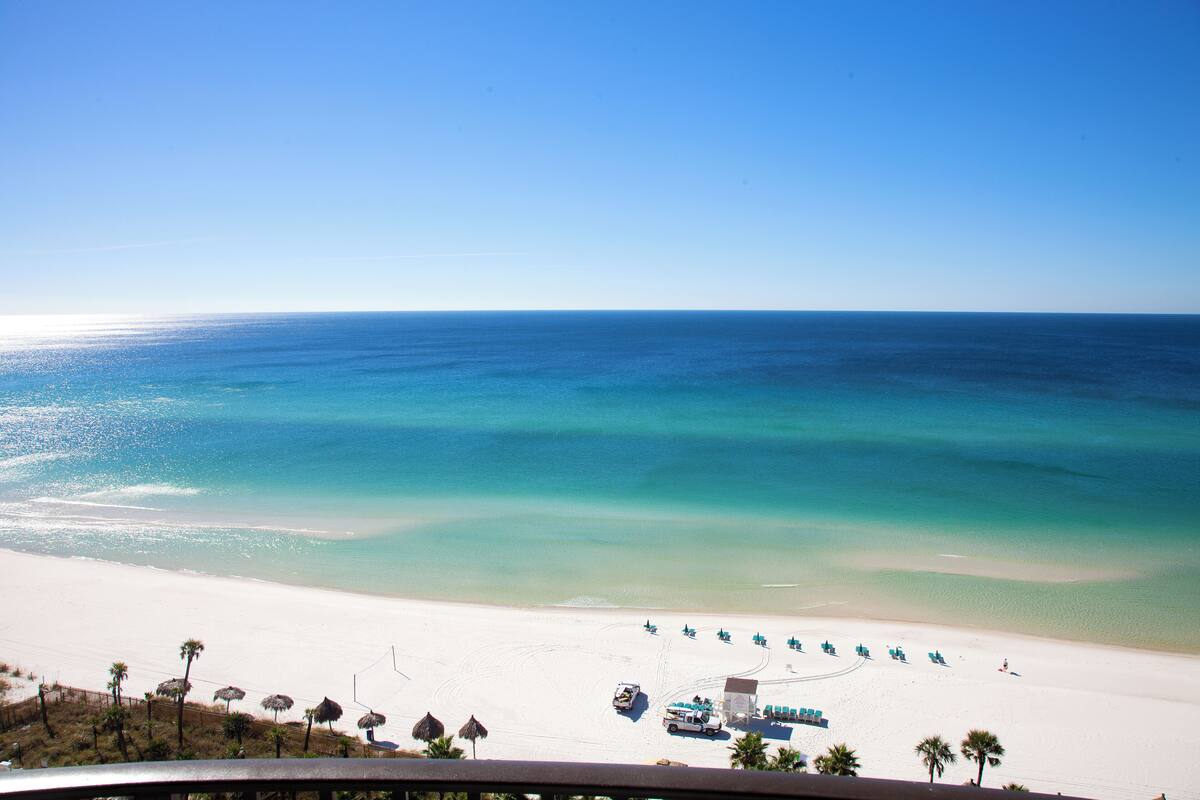 A beach with clear turquoise water, white sand, and a few palm trees. There are umbrellas and lounge chairs along the shore.