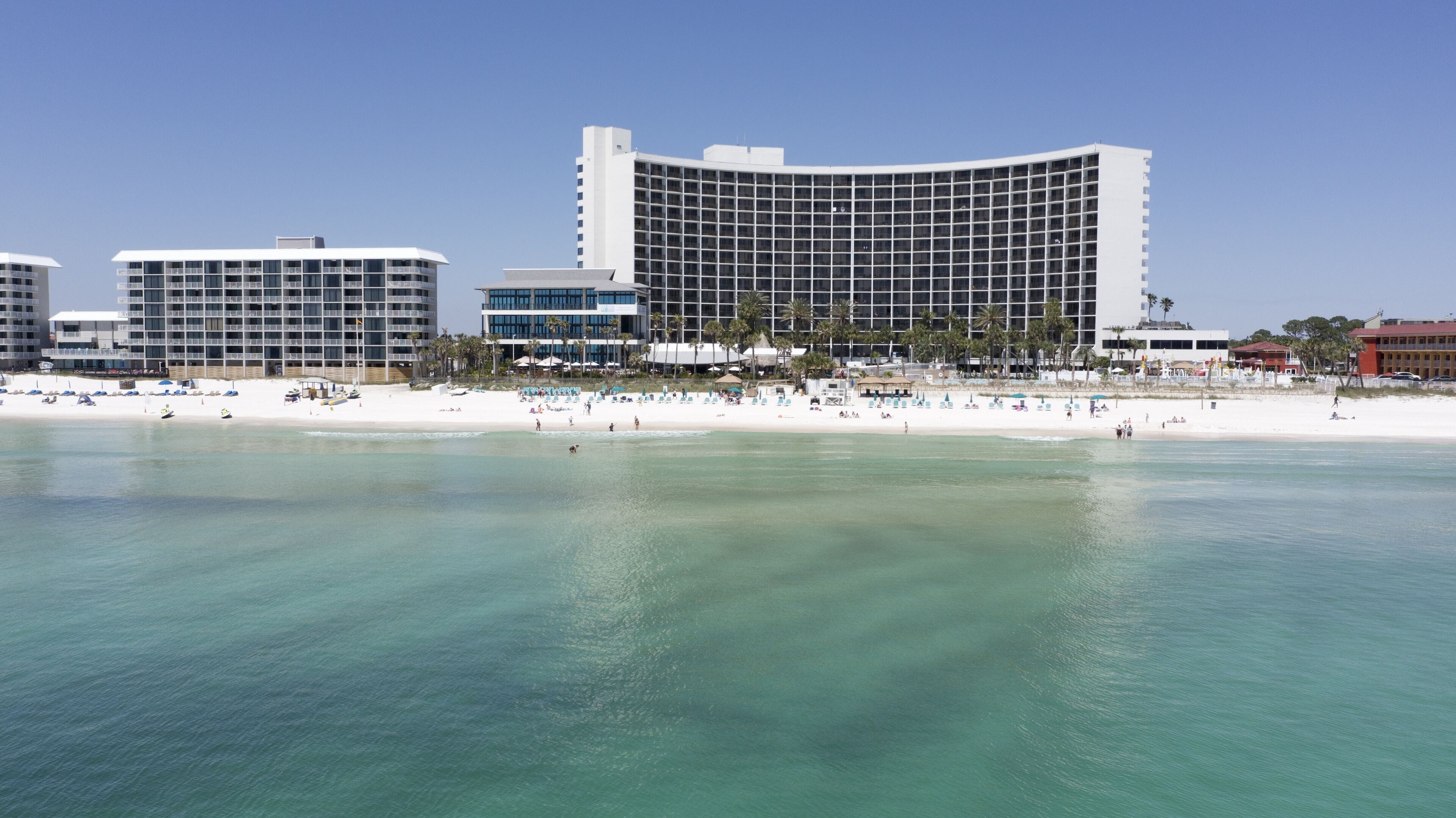 Un hotel frente a la playa con fachada curva, varios balcones y una playa arenosa al frente.