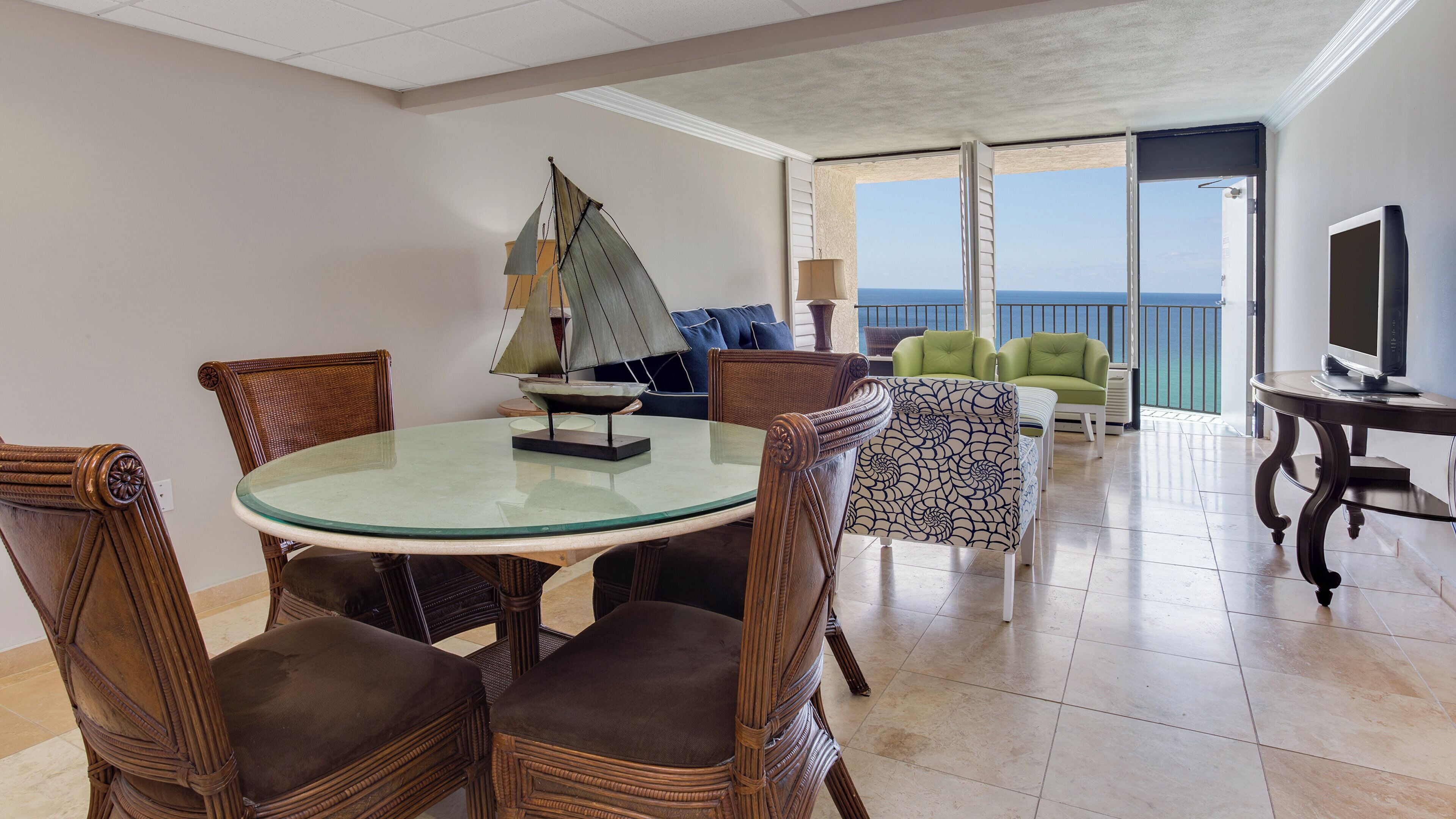 Una habitación con una mesa redonda de cristal, sillas de mimbre, un modelo de velero, un sofá con cojines azules, un sillón verde, un televisor en un mueble bajo y un balcón con vistas.