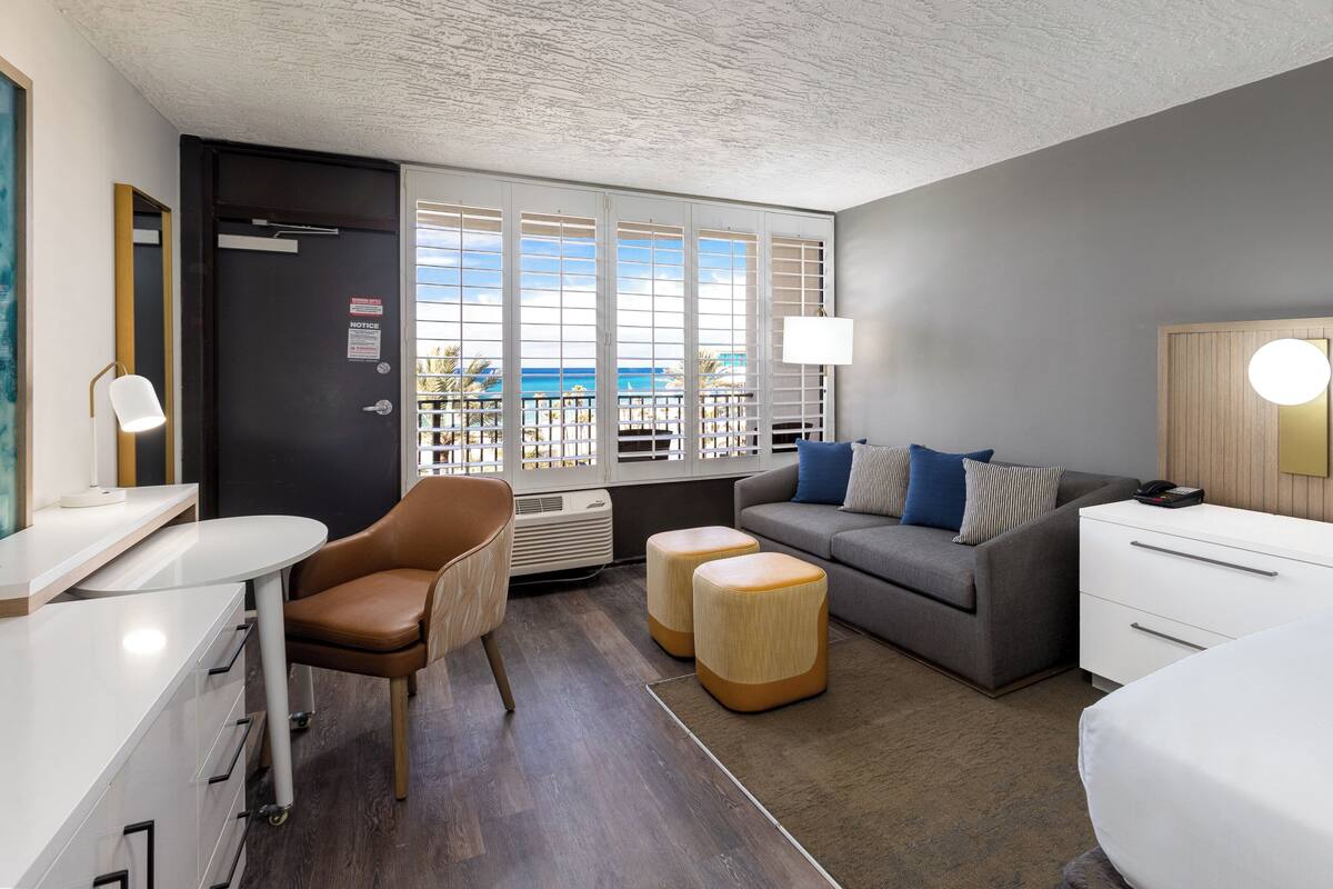A modern hotel room with a grey sofa, a bed, a desk, and a view of the ocean through large windows.