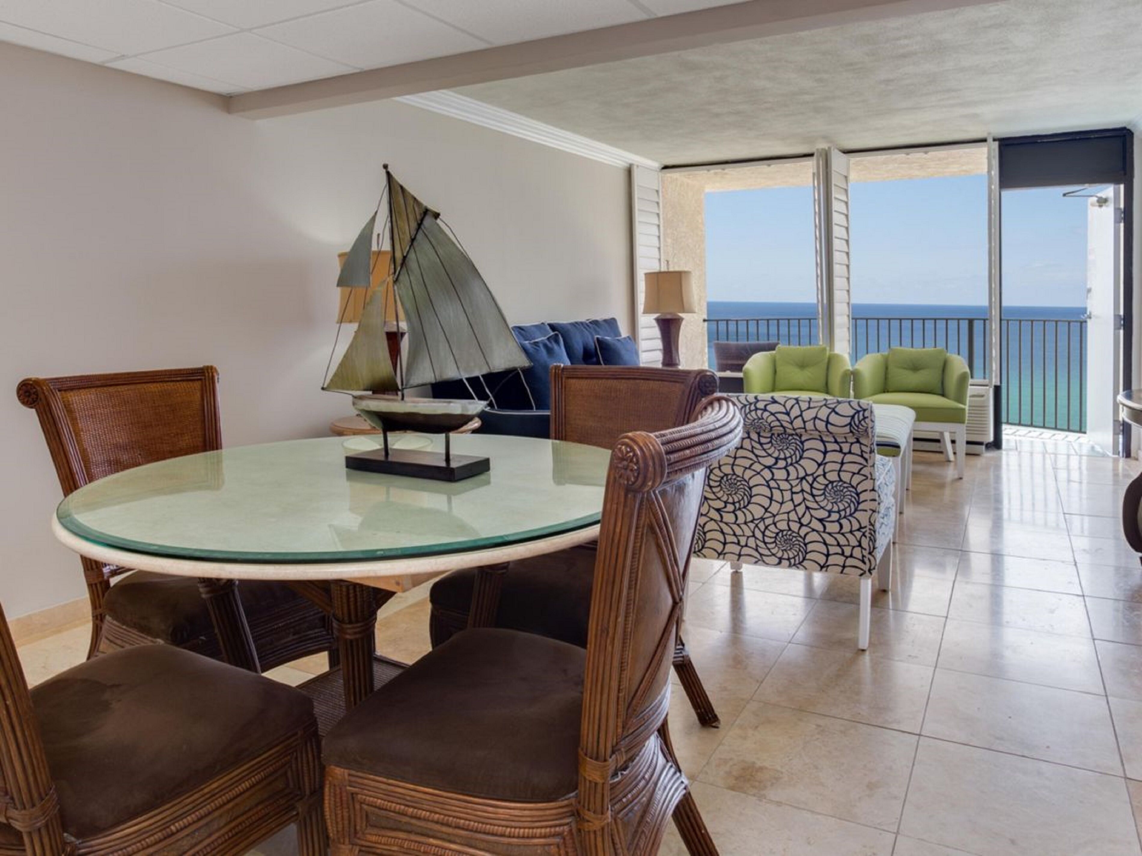 A dining area with a glass table, wicker chairs, and a sailboat model. There is a sofa with patterned cushions and a view of the ocean through large windows.