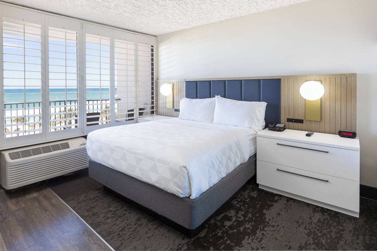 Habitación de hotel con cama, mesita de noche y vista al mar a través de amplias ventanas.