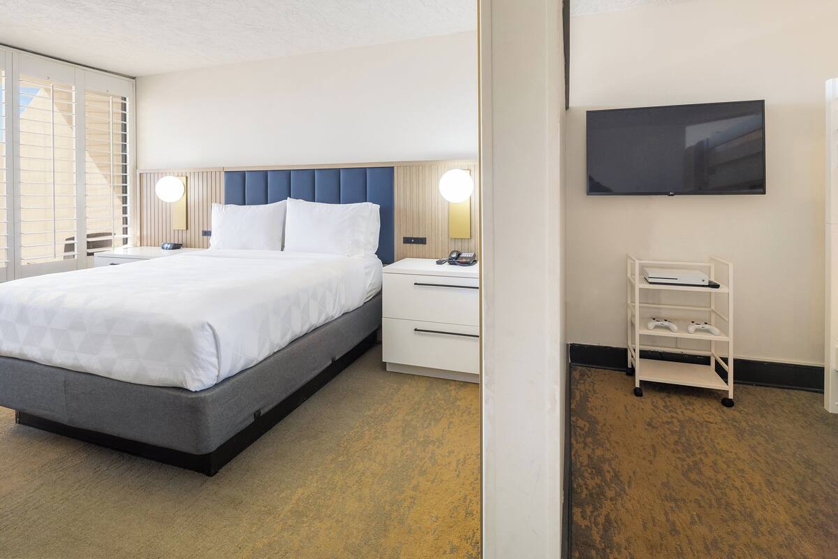 A hotel room with a bed, bedside tables, a TV, and a small shelf with items.
