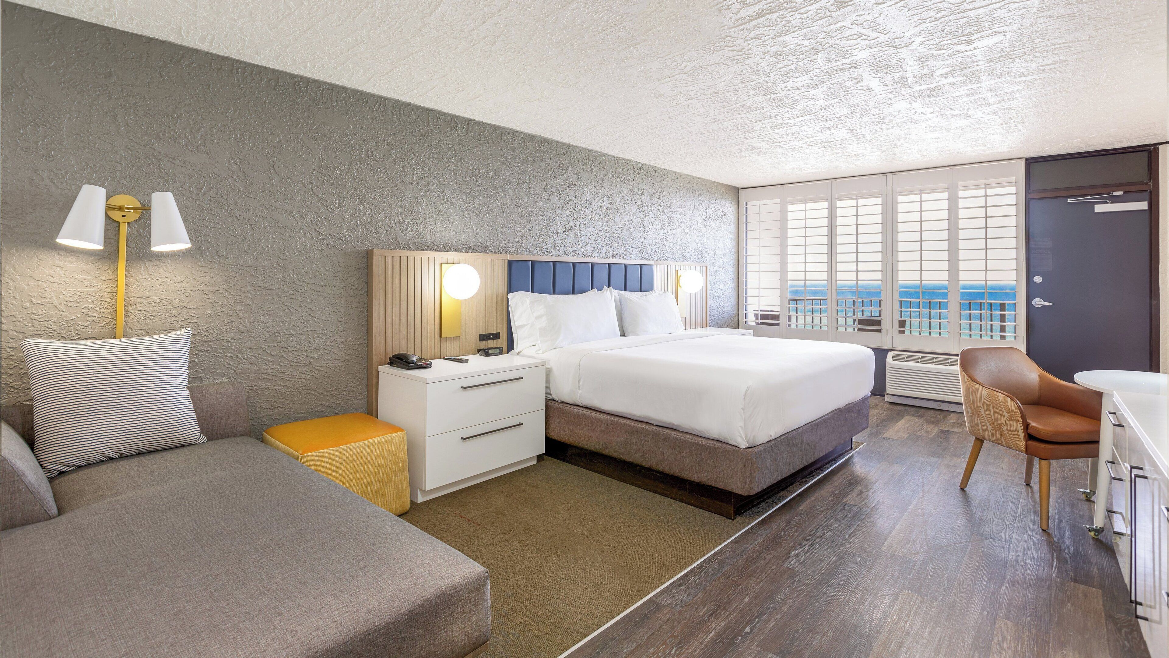 A modern hotel room with a large bed, a bedside table, a chair, and a view of the sea through large windows.