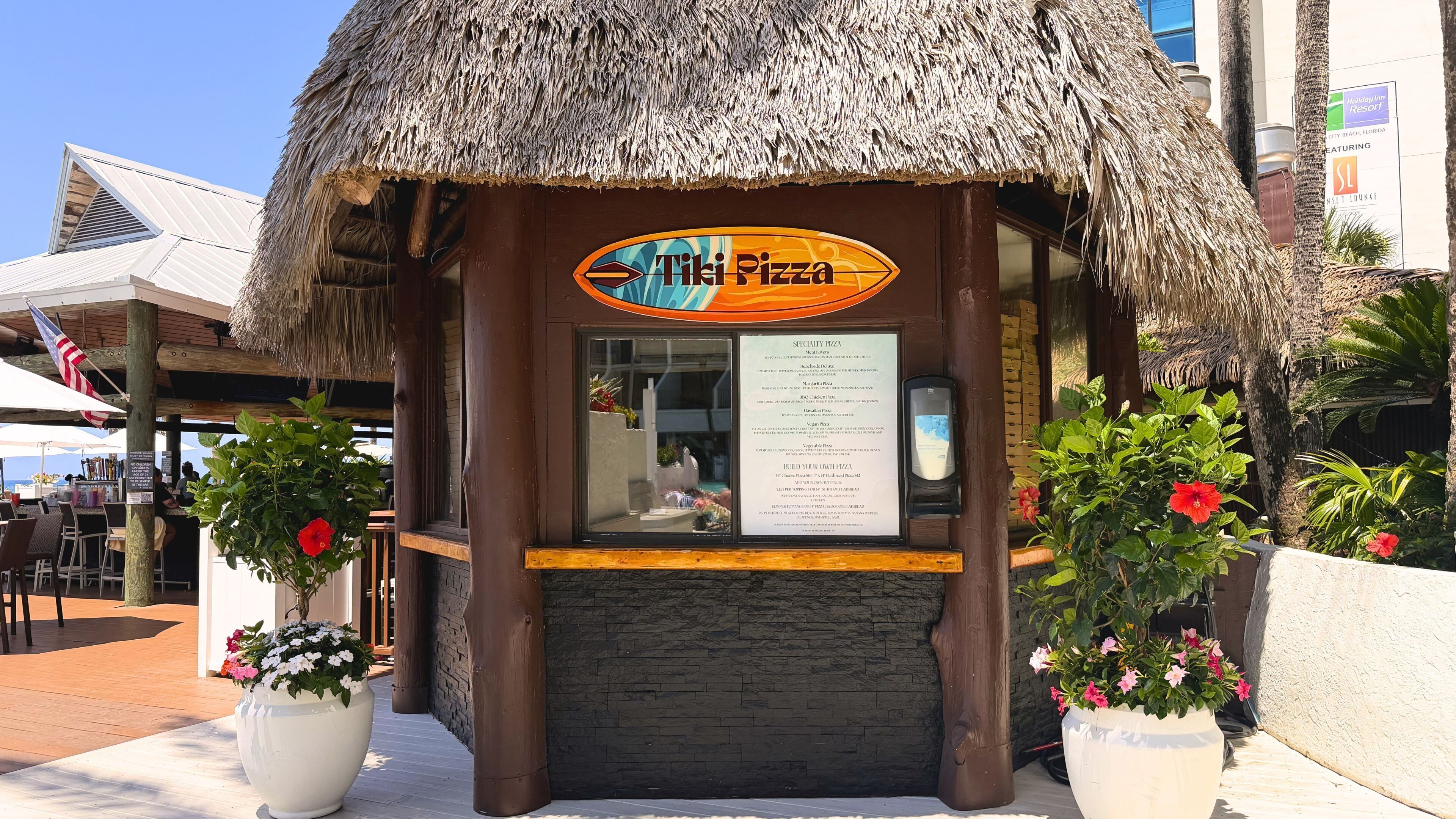 Un restaurante de temática tiki con techo de paja, un tablero de menú y plantas en macetas.