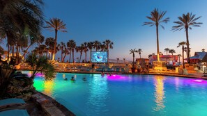 Un área de piscina con palmeras, una pantalla grande y sillones de descanso.