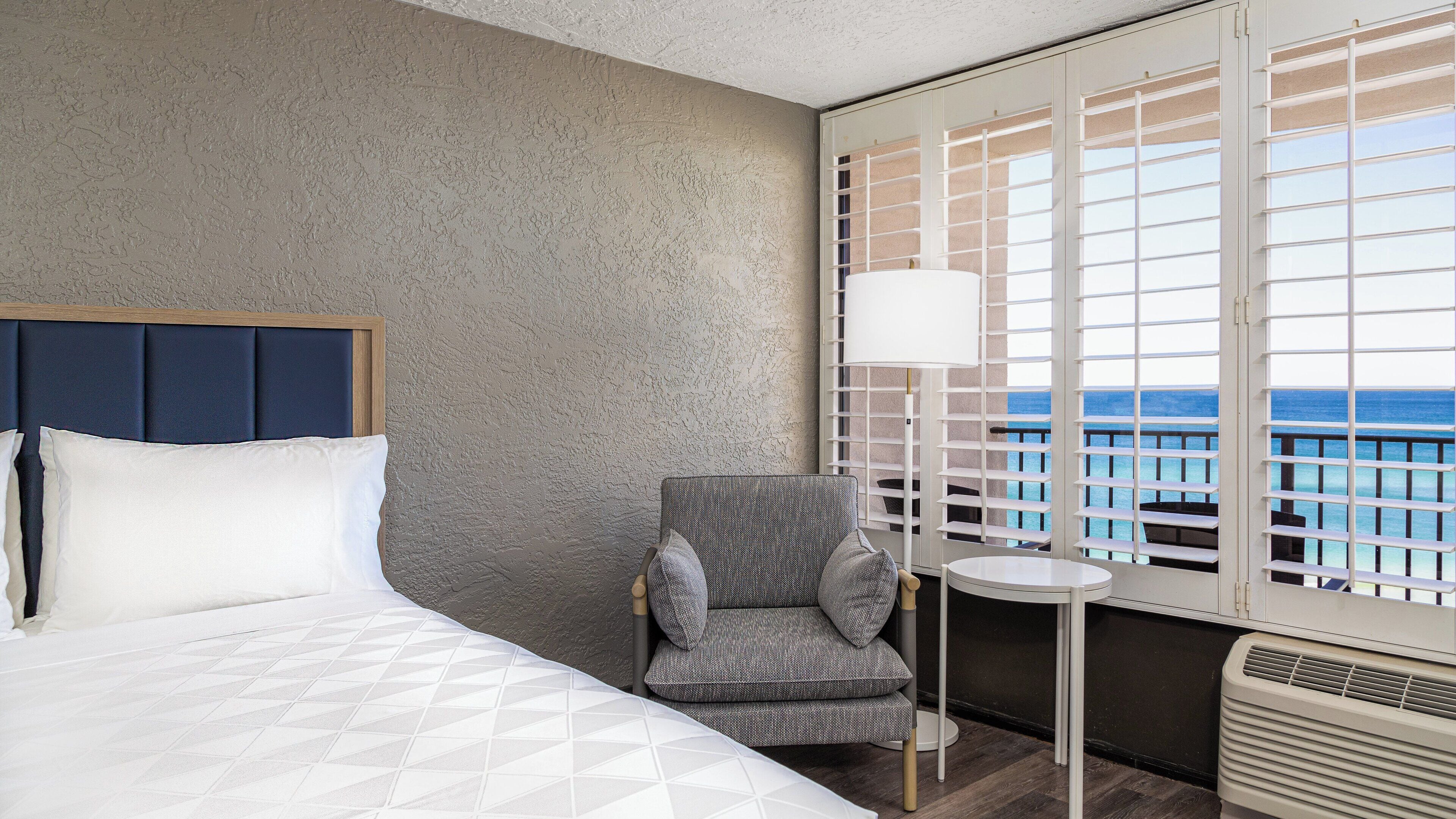 Habitación de hotel con cama, una silla, una mesita y vista al océano a través de una ventana con persianas.