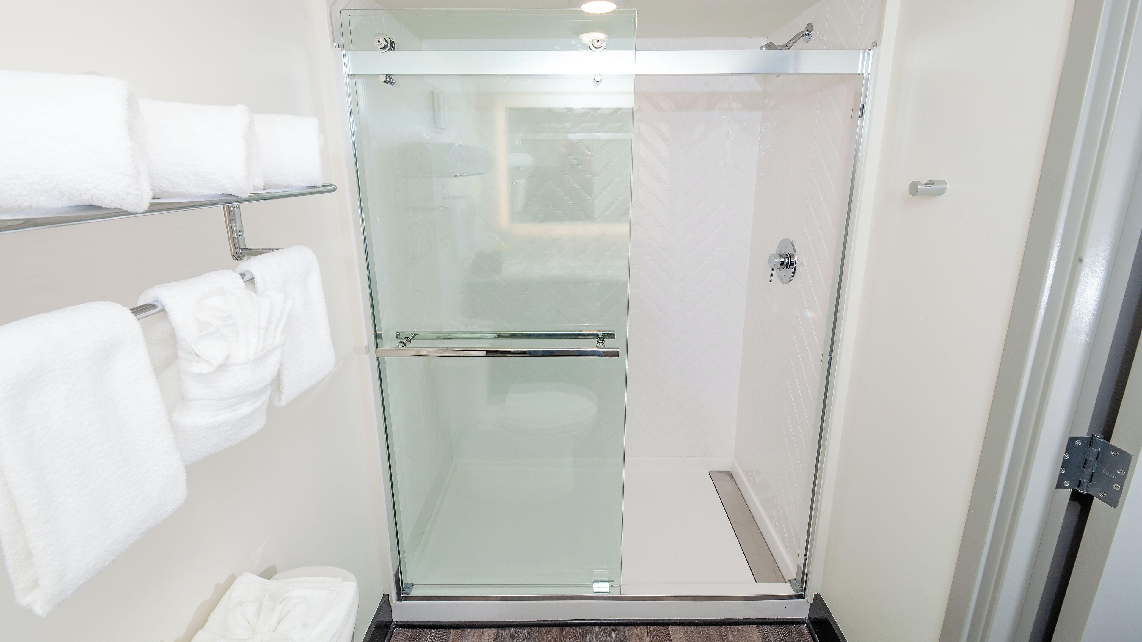 A glass-enclosed shower with a white tiled wall, a towel rack with multiple white towels, and a visible shower control panel.
