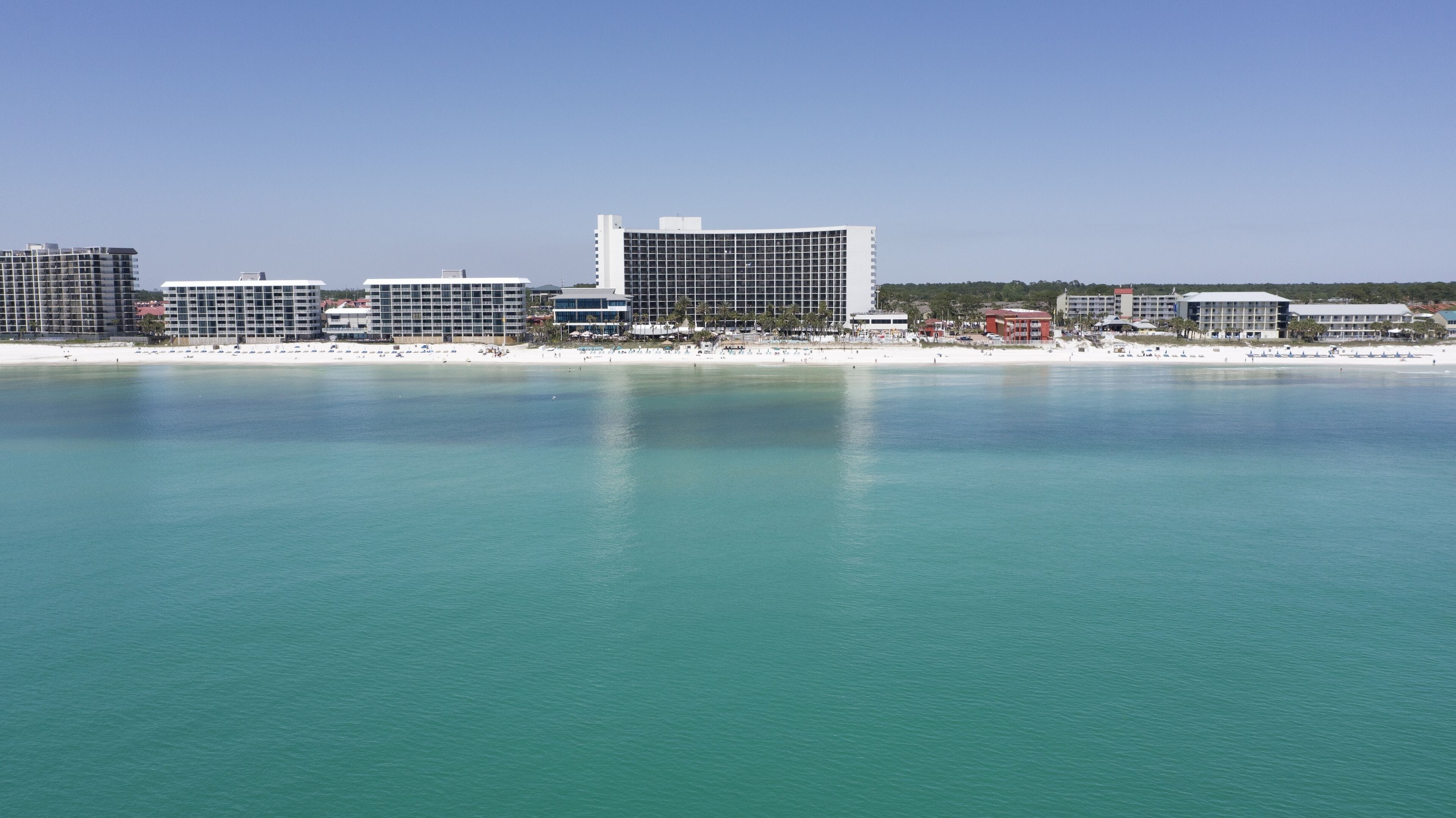 A coastal cityscape with a prominent hotel featuring a modern design, situated near a beach with clear turquoise waters.