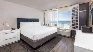 A modern hotel room with a bed, bedside tables, a desk, a chair, a window with a view of the beach, and a balcony with a railing.