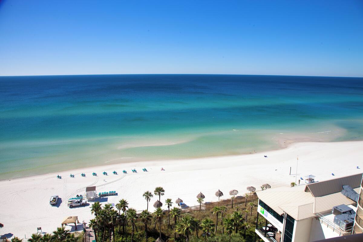 A beach view with a clear sky, a sandy beach, and a building with a flat roof and balconies.