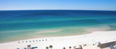 A beach view with a clear sky, a sandy beach, and a building with a flat roof and balconies.