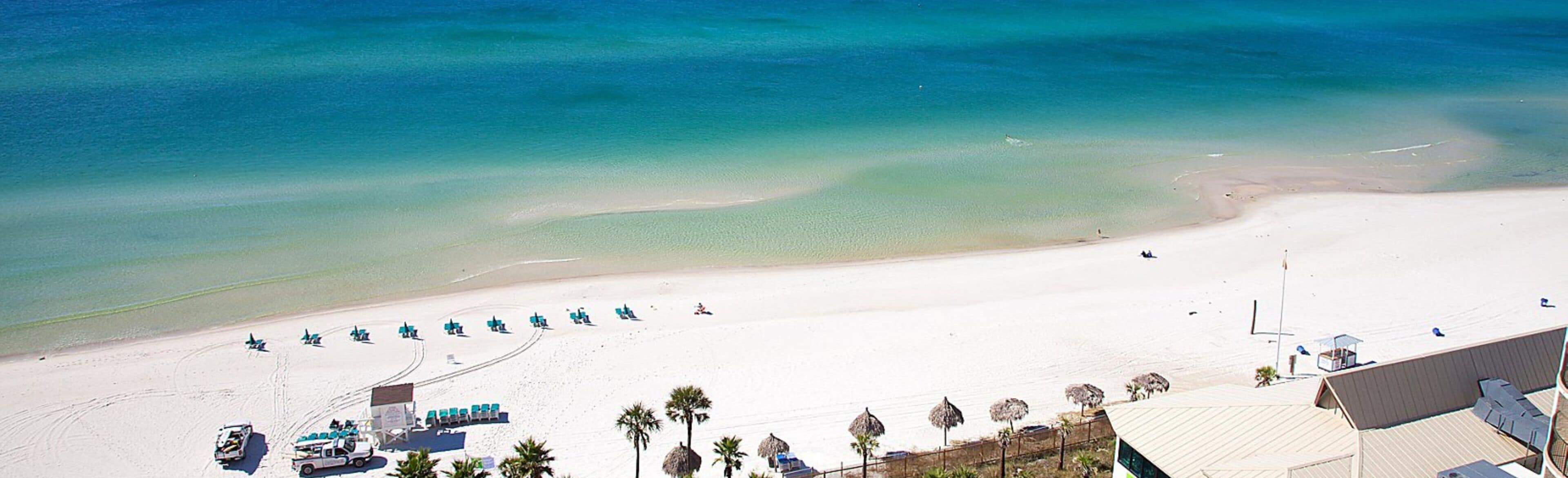Vista aérea de una playa con una fila de sillas y sombrillas azules, una torre de salvavidas y un edificio de techo inclinado.
