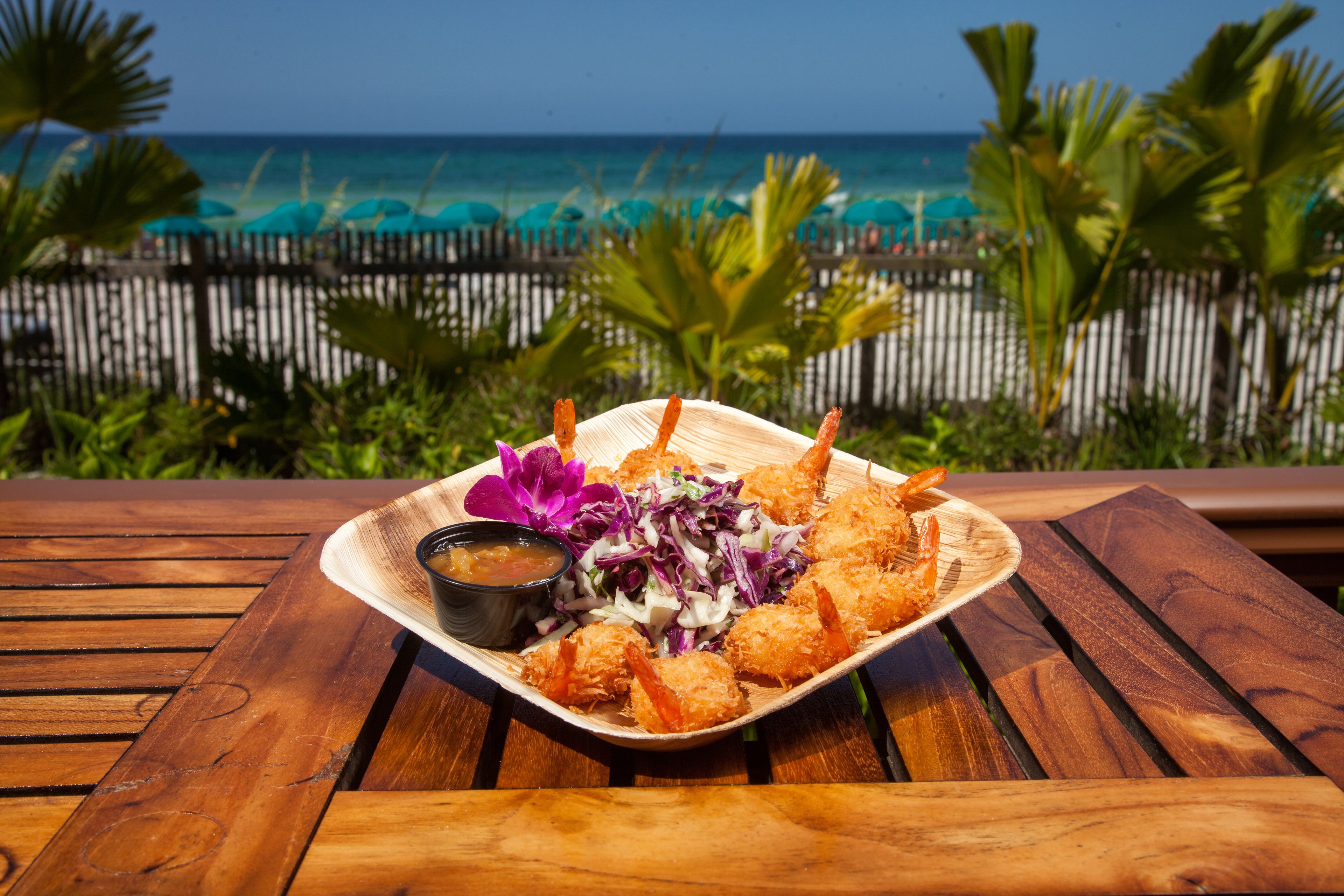A wooden table with a plate of fried shrimp and salad, set against a view of the ocean and beach.