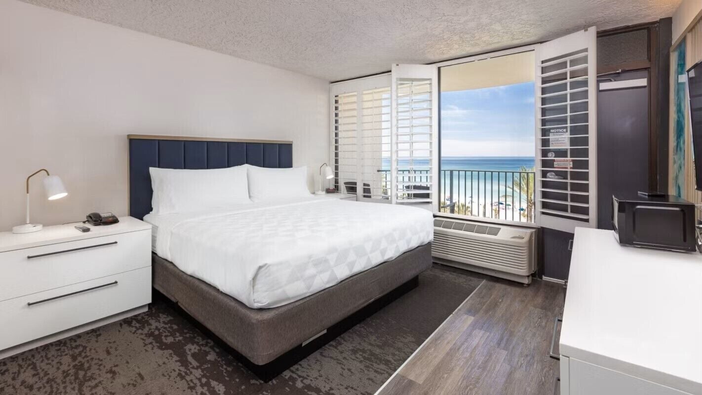 A modern hotel room with a bed, bedside table, lamp, and a view of the ocean through a balcony.
