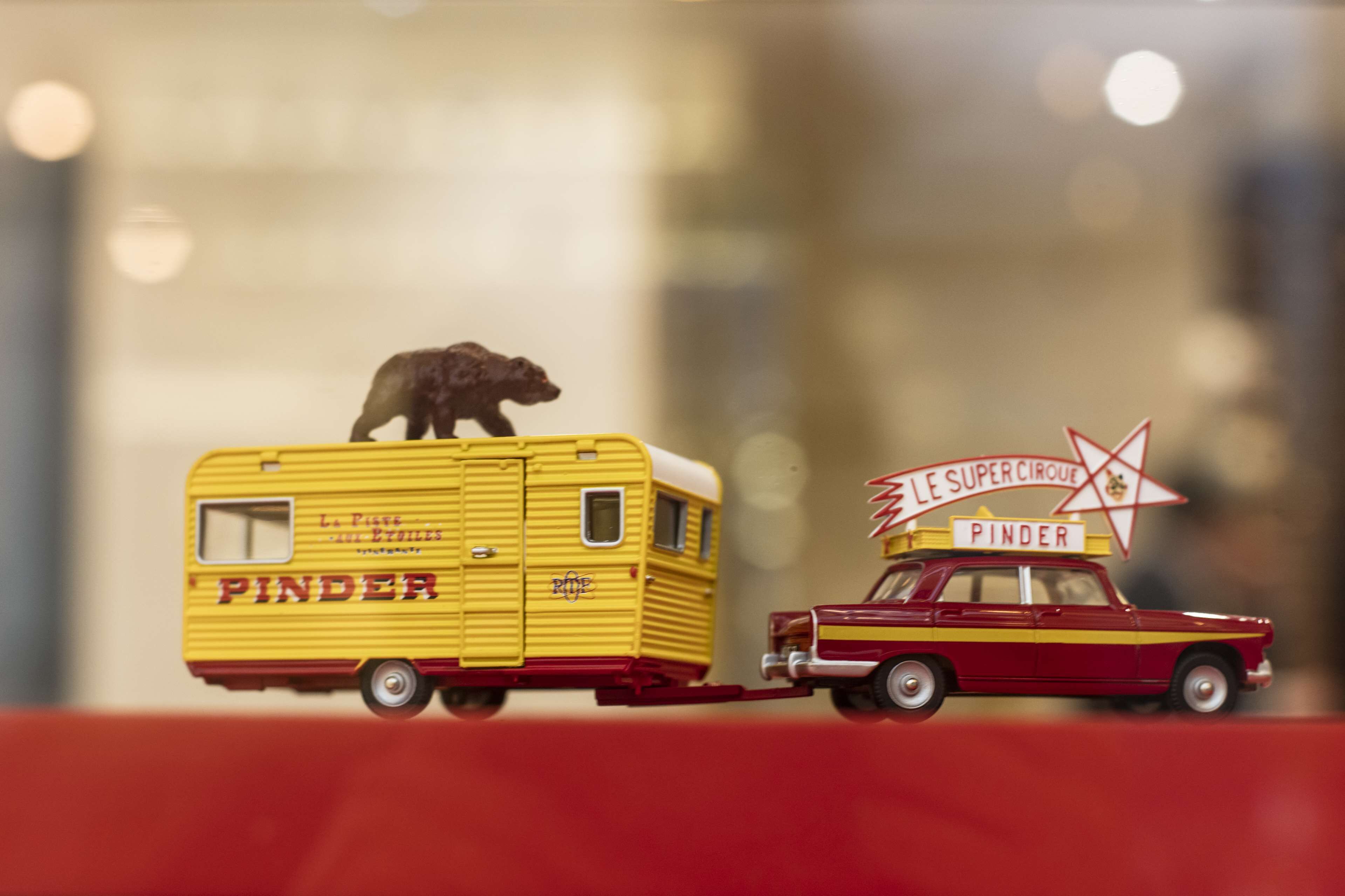 A yellow trailer with "PINDER" written on it, a red car with "LE SUPER CIRQUE" and "PINDER" on the roof, and a bear on the trailer.