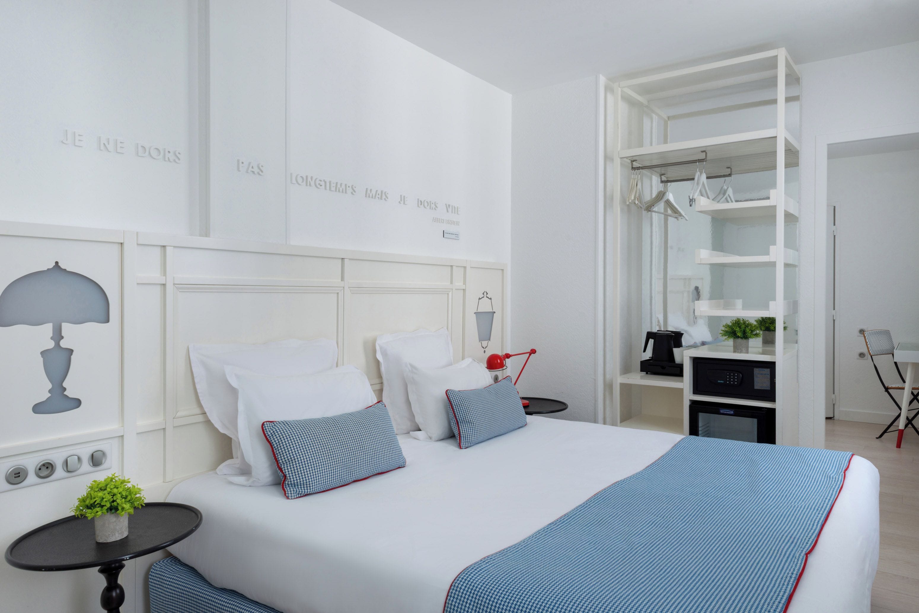 A modern hotel room with a neatly made bed, a side table with a lamp, a small potted plant, and a built-in wardrobe with hanging space.