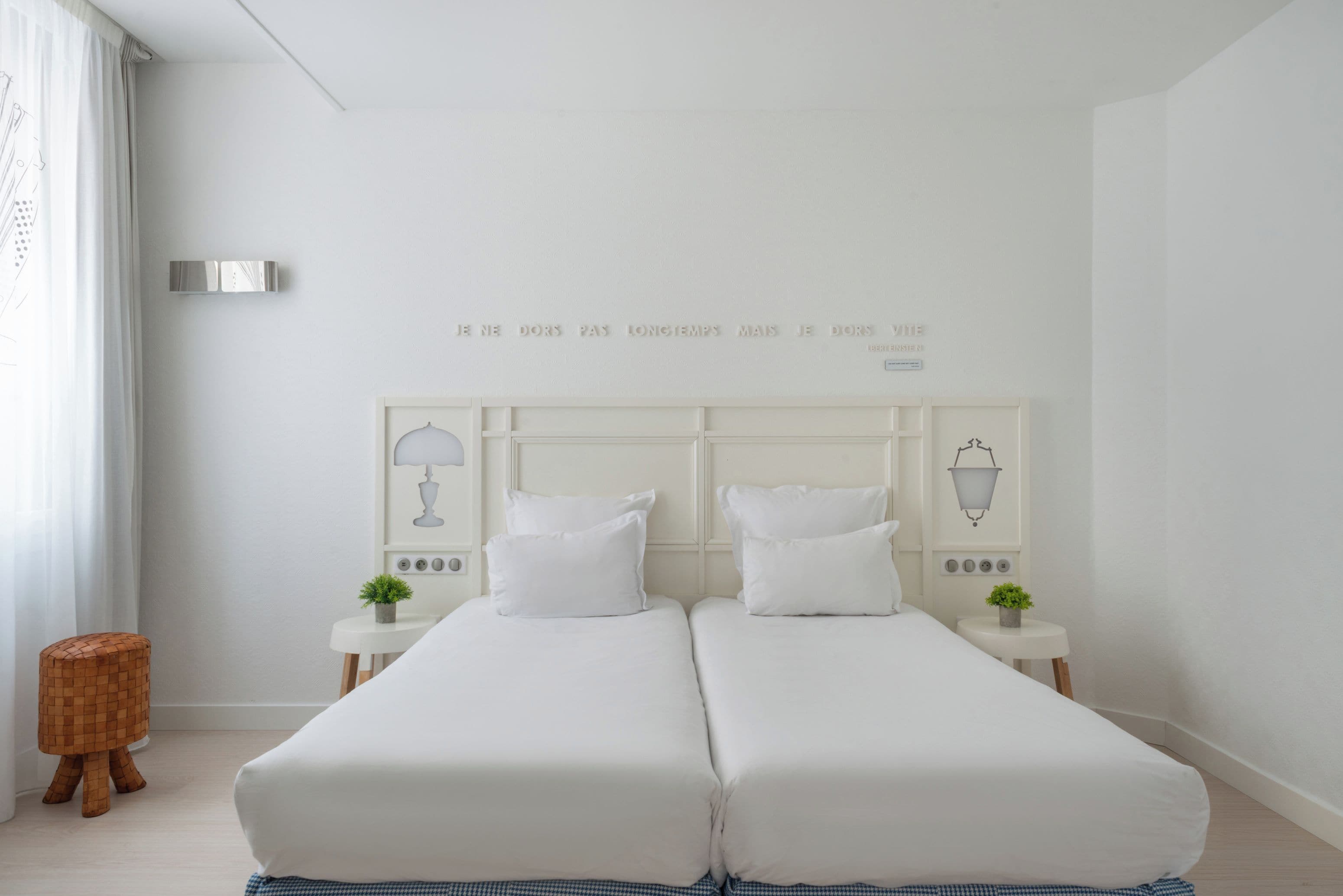 A double bed with white bedding, two bedside tables with lamps, and a wall with a French phrase.