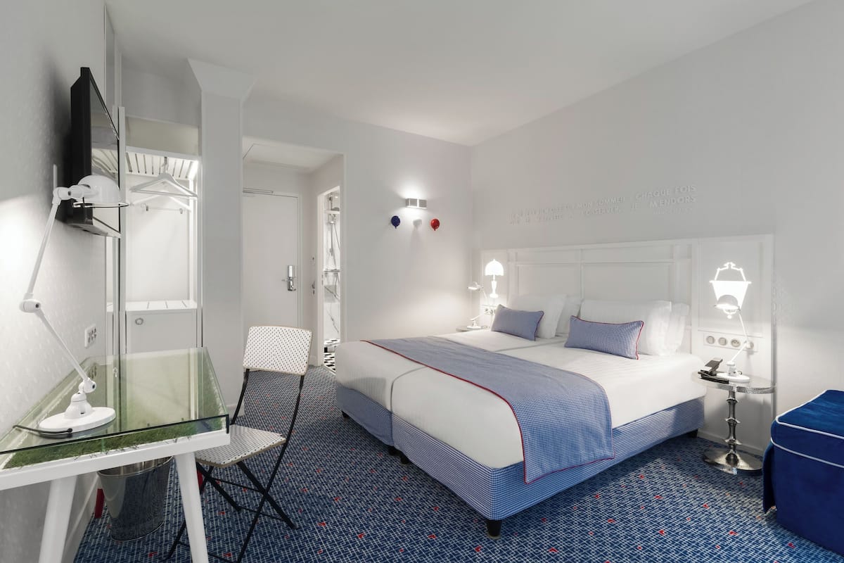 Une chambre d’hôtel avec un grand lit, un bureau en verre avec une chaise, une table de chevet avec une lampe et une armoire.