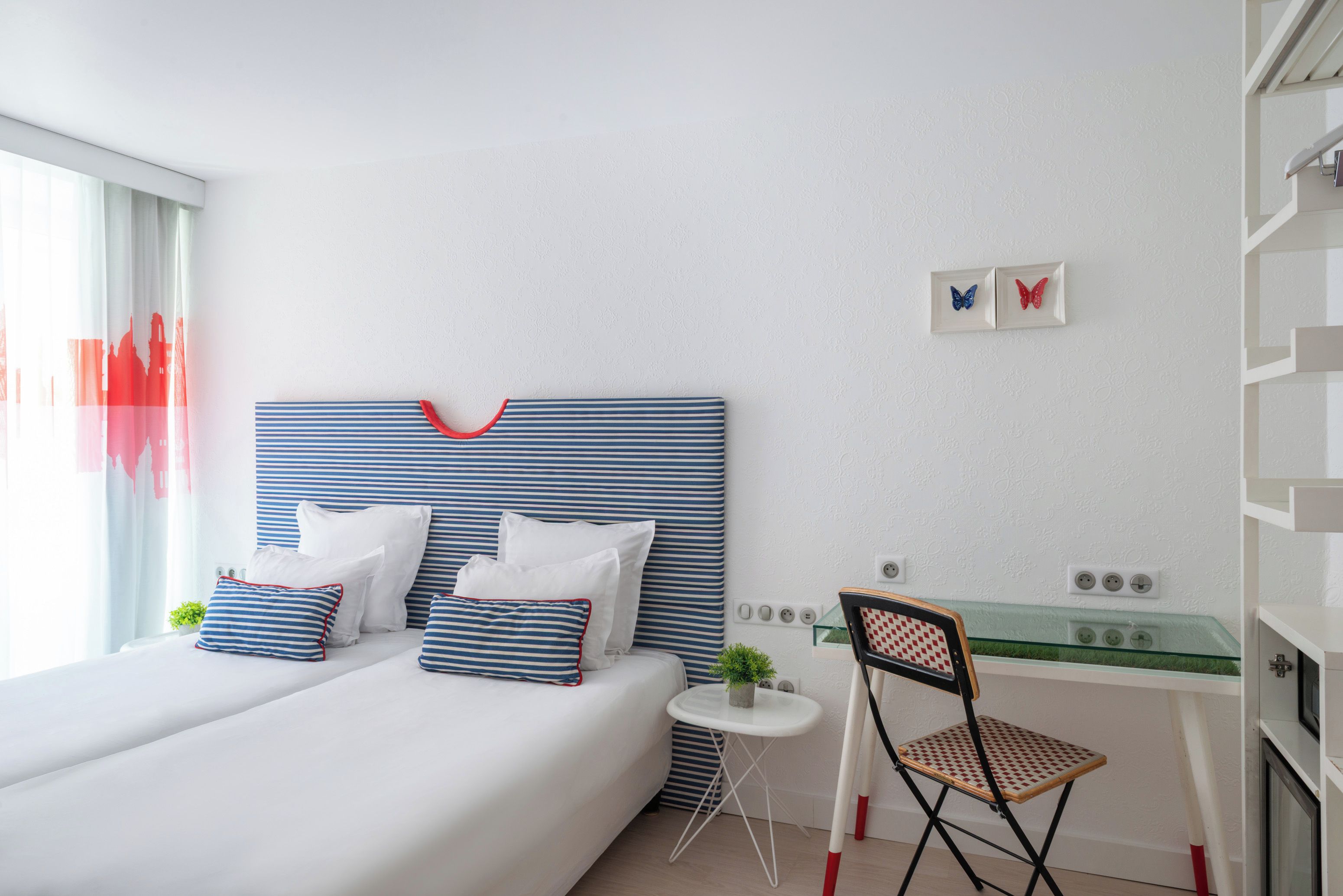A hotel room with a bed, pillows, a small table, a chair, and a wall-mounted power outlet.