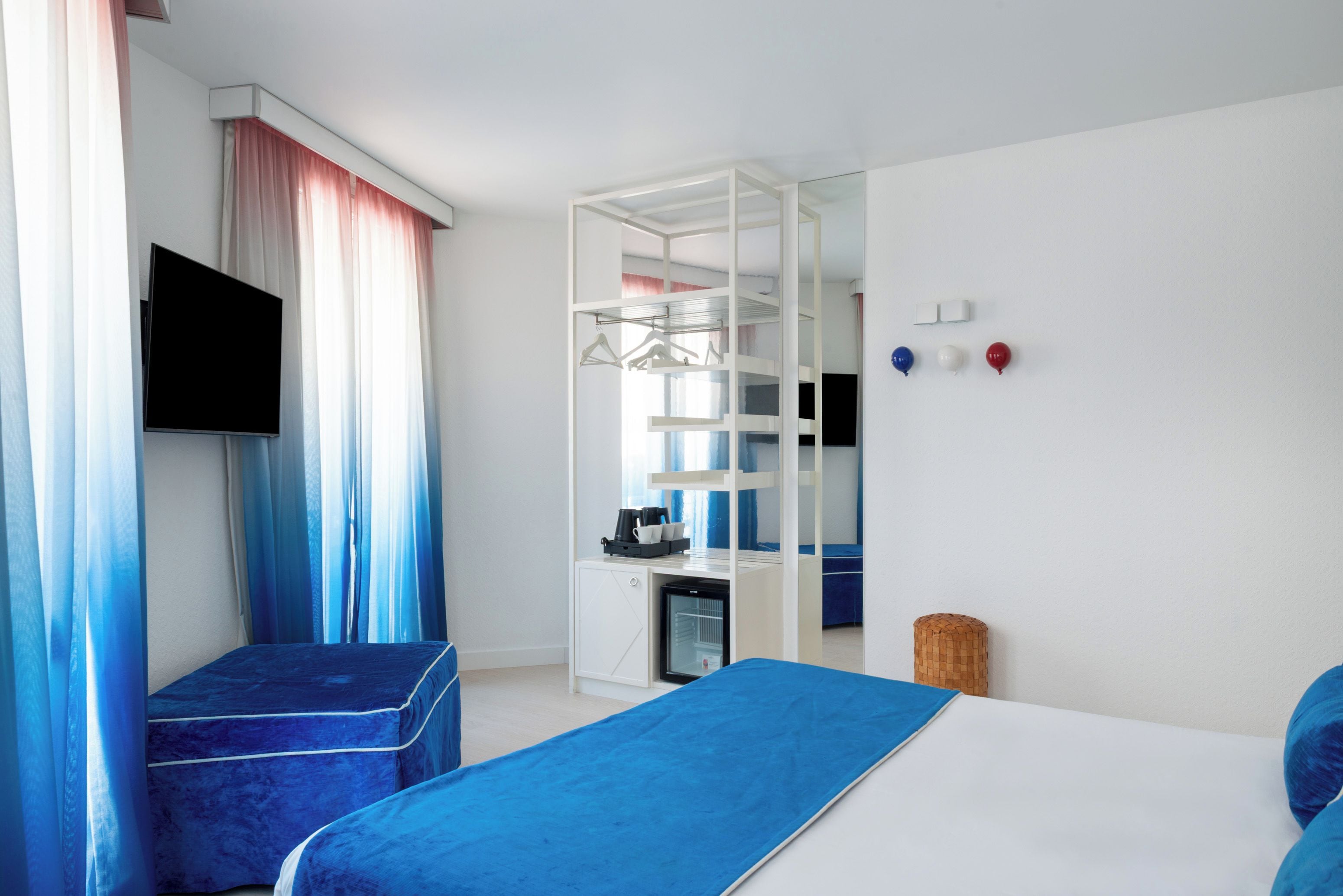 A modern hotel room with a bed, a seating area, a desk with a computer, a wardrobe, and a television.