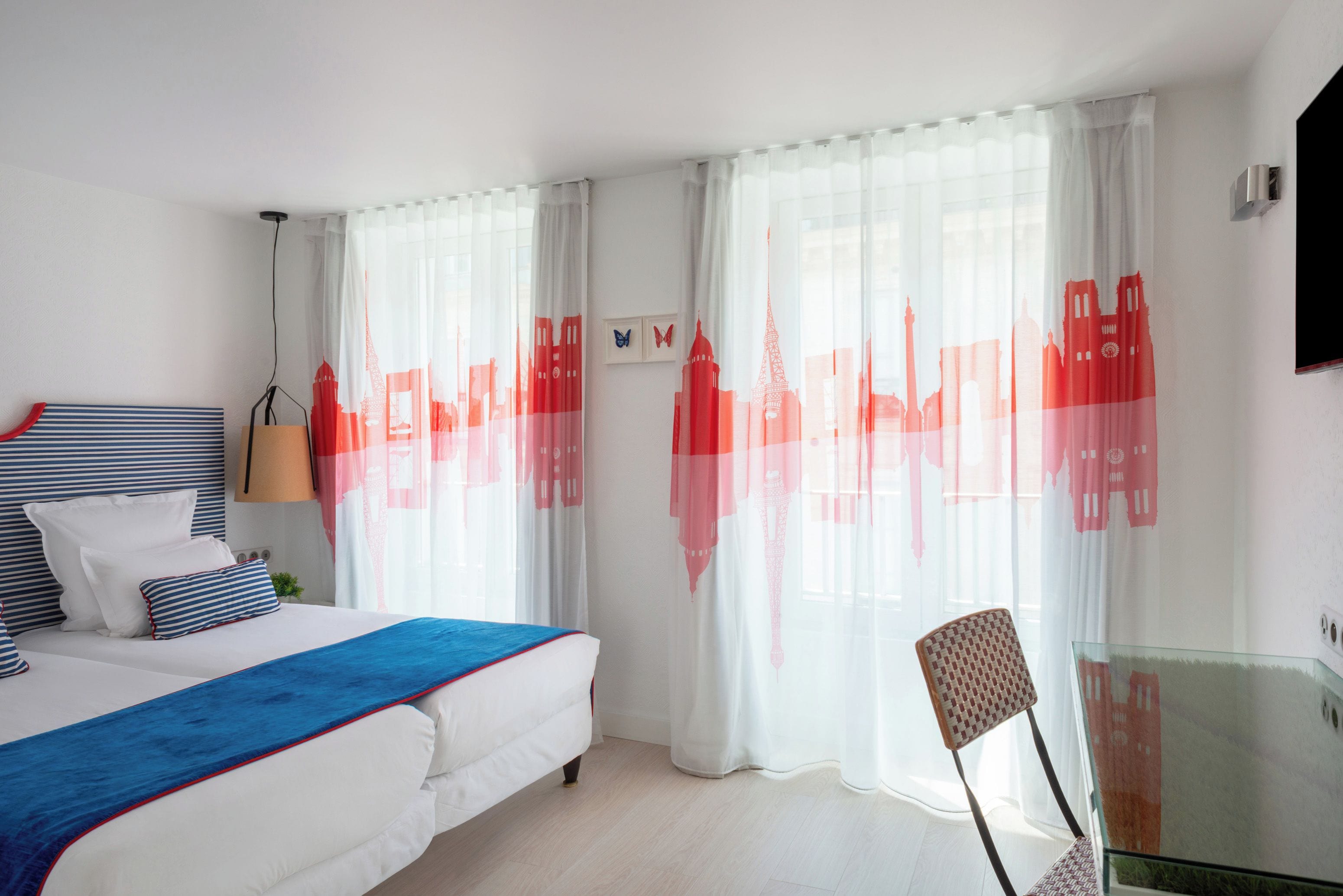 Une chambre d’hôtel avec un lit, une chaise, une table en verre et des rideaux ornés d’un motif représentant un paysage urbain.