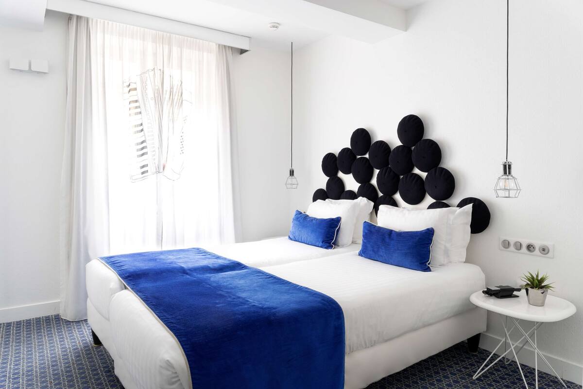 Une chambre d’hôtel moderne avec un grand lit, une petite table de chevet agrémentée d’une plante et une tête de lit originale en noir et blanc.