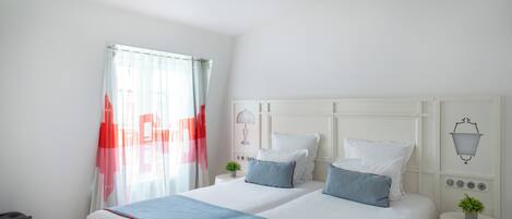 A hotel room with two beds, a small table, a chair, a window with curtains, and a wall-mounted lamp.