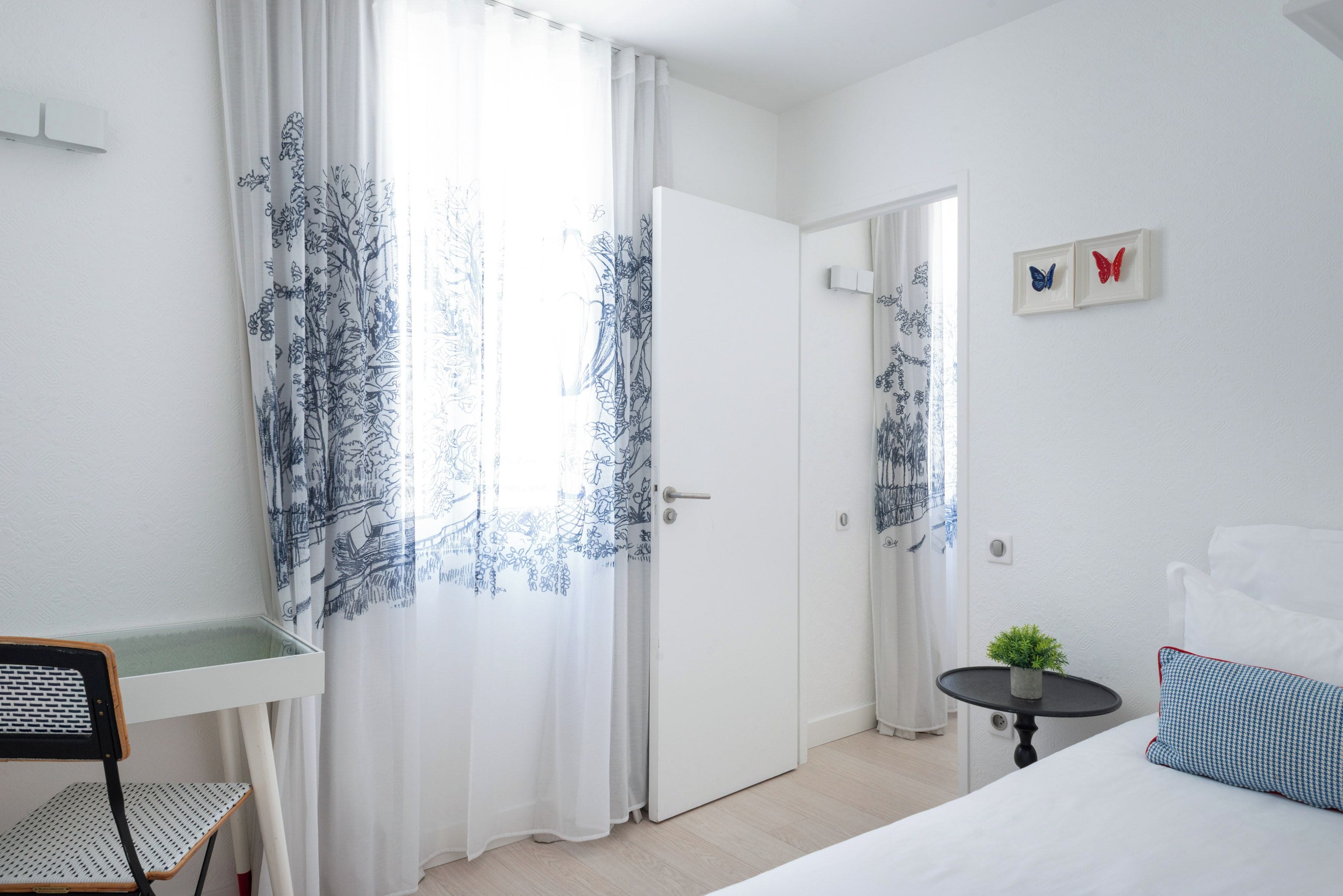 A hotel room with a bed, a desk, a chair, a small table with a plant, and a door leading to another room.