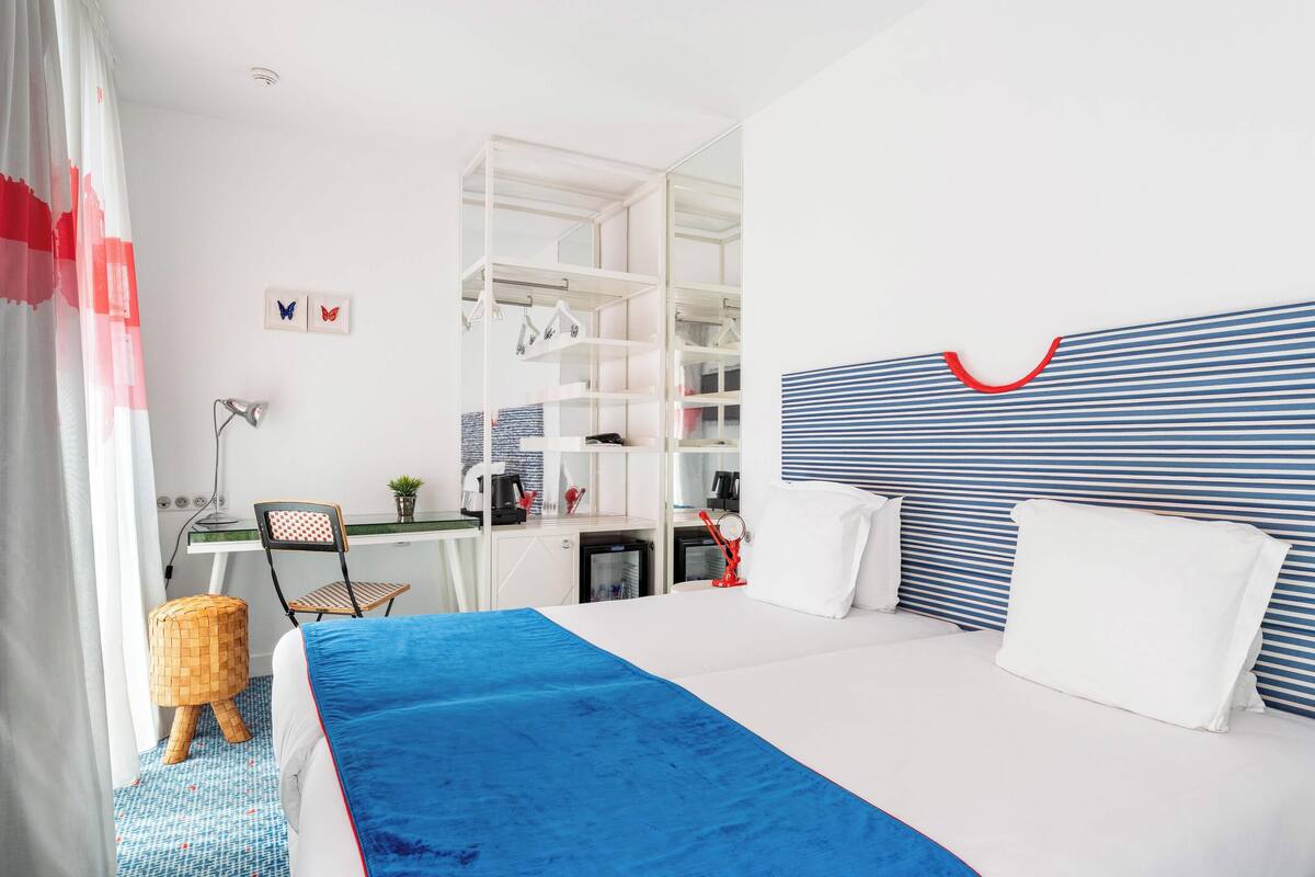 Une chambre d’hôtel comprenant un lit, un bureau, une chaise, une armoire et un petit tabouret.