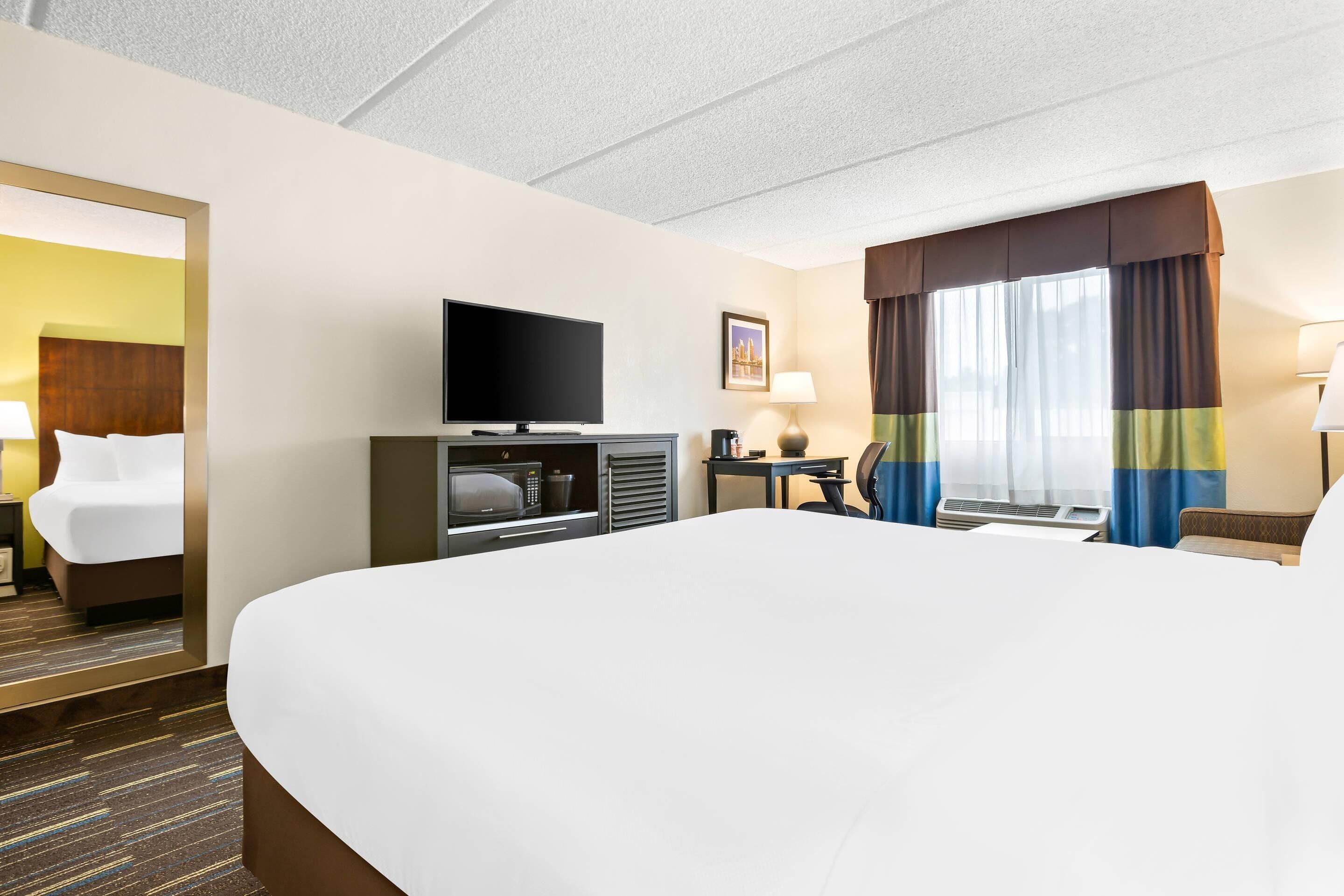 Ein Hotelzimmer mit einem großen Bett, einem Flachbildfernseher, einem Schreibtisch mit Stuhl, einer Mikrowelle und einem Fenster mit Vorhängen.
