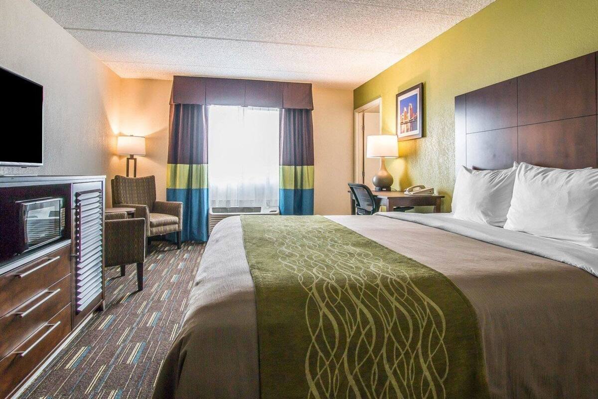 A hotel room with a bed, a desk with a lamp, a chair, a television, and a window with curtains.