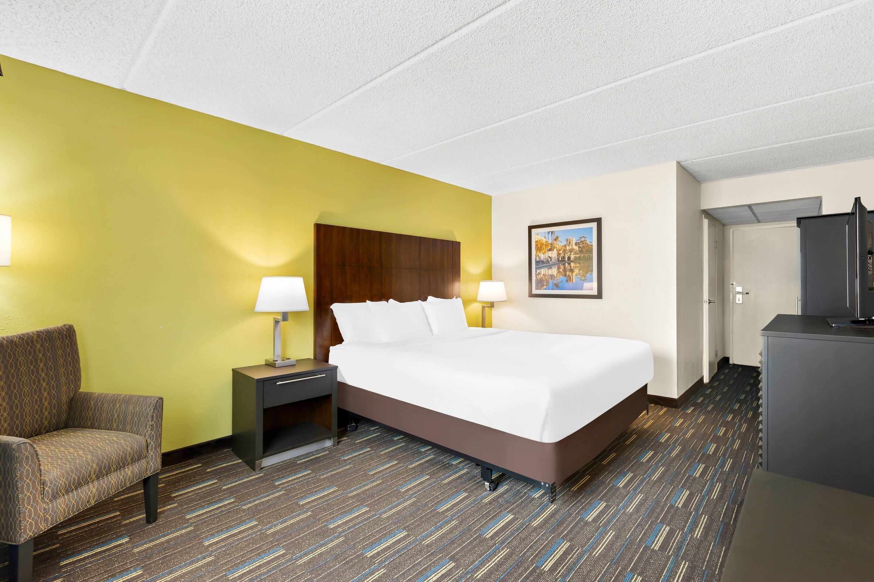 A hotel room with a bed, a chair, a desk, a lamp, a painting, and a television.