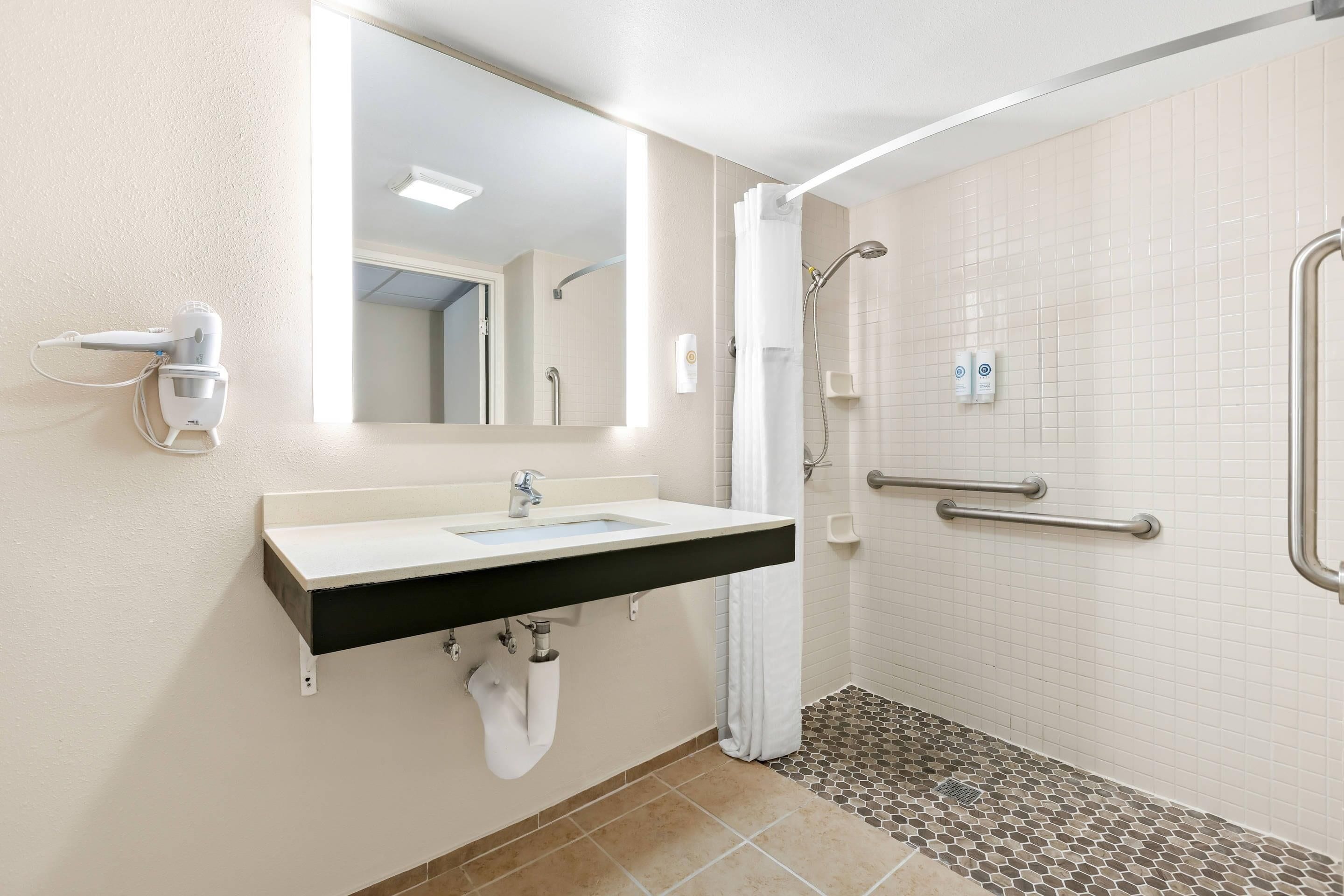 A bathroom with a sink, mirror, towel rack, shower with grab bars, and a tiled floor.