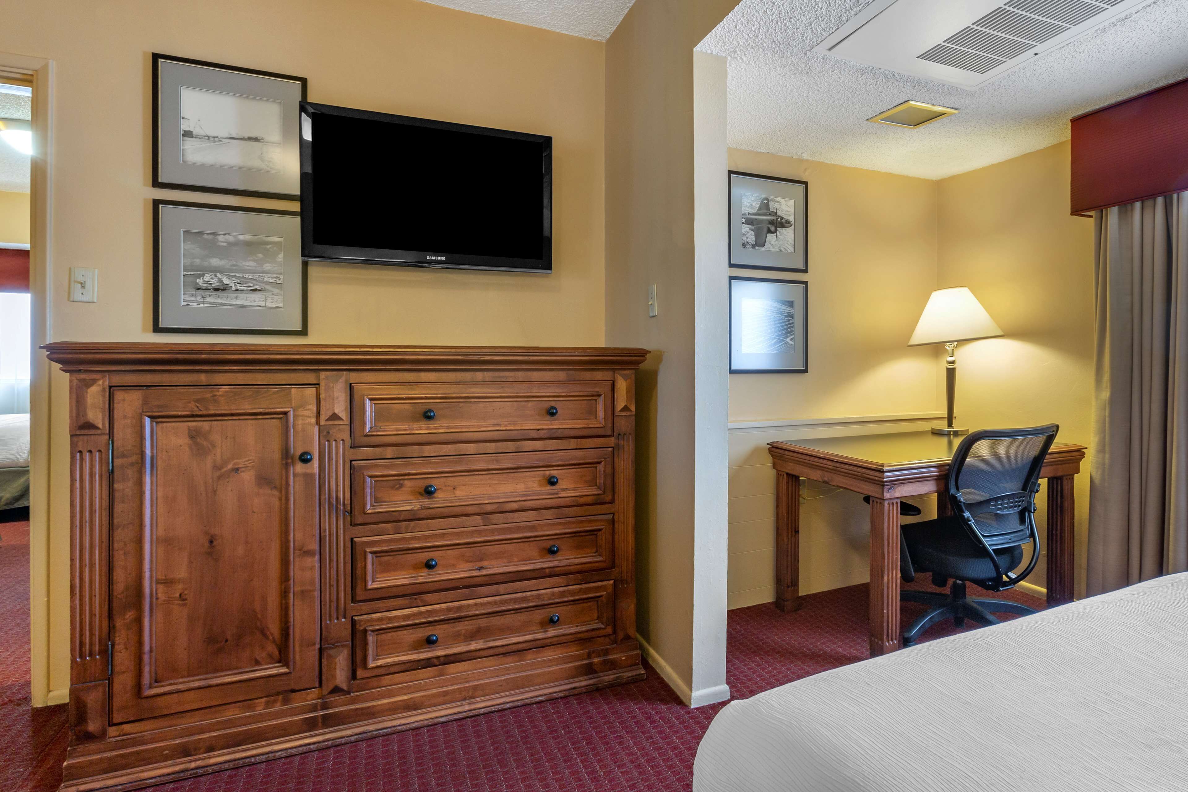 A hotel room with a wooden dresser, a flat-screen TV, a desk with a chair, and a lamp.