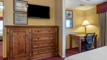 A hotel room with a wooden dresser, a flat-screen TV, a desk with a chair, and a lamp.