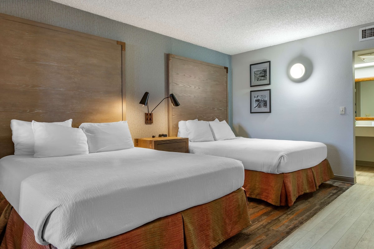 A hotel room with two beds, a wooden headboard, a nightstand, a wall-mounted lamp, and two framed pictures.