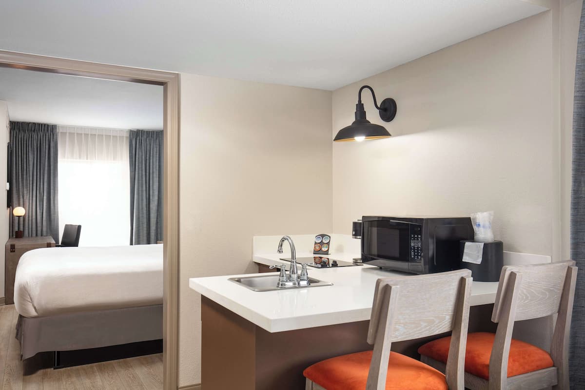 A hotel room with a bed, a small kitchenette with a microwave, a sink, and a chair with an orange cushion.