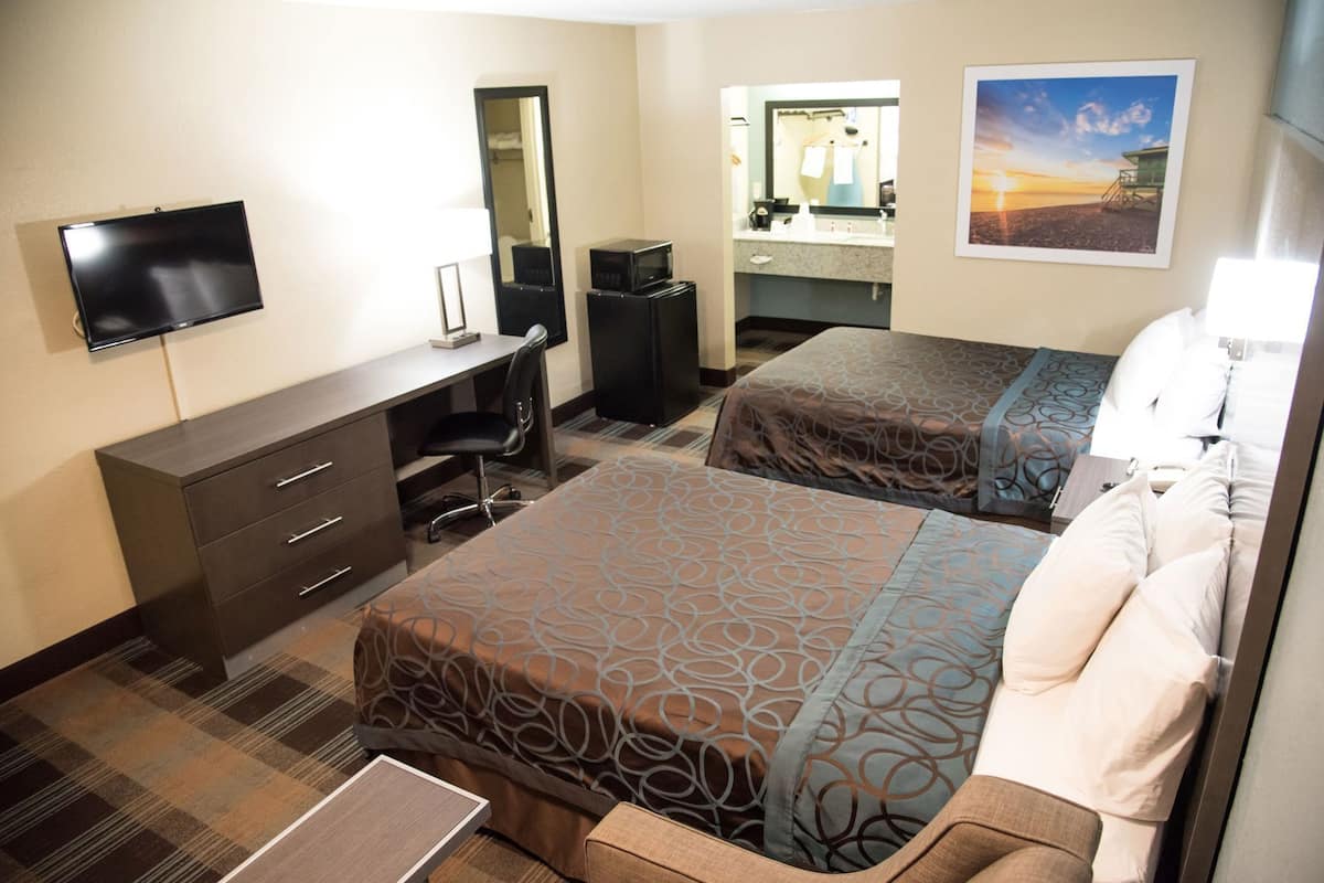 Habitación de hotel con dos camas, un escritorio, una silla, televisión, espejo y baño visible a través de una puerta abierta.