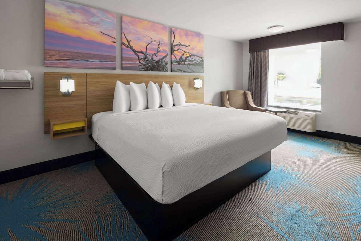 Habitación de hotel con una cama grande, cabecera de madera, mesita de noche, una silla, ventana con cortinas y dos lámparas de pared.