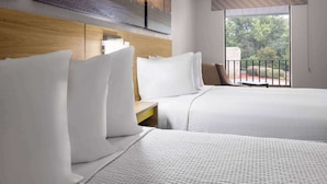 A hotel room with two beds, a wooden headboard, a window with a view of greenery, and a balcony with a railing.