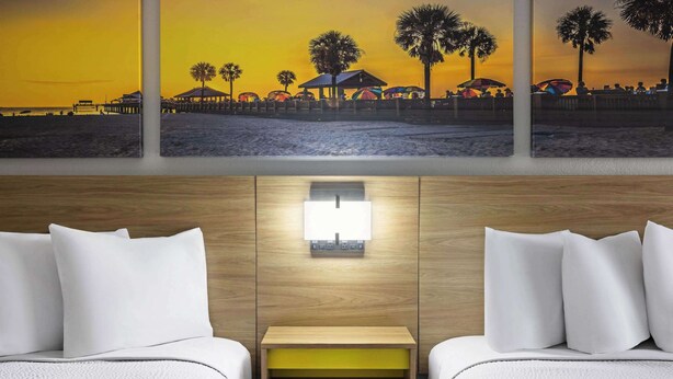 A hotel room with two beds, a nightstand, a wall-mounted lamp, and a beach scene painting above the beds.