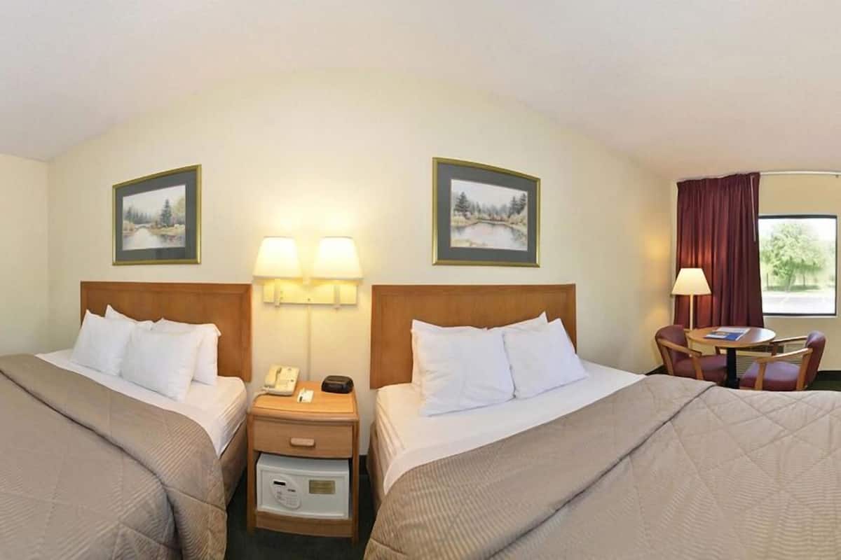 A hotel room with two beds, a desk with a lamp, a chair, and a window with curtains.
